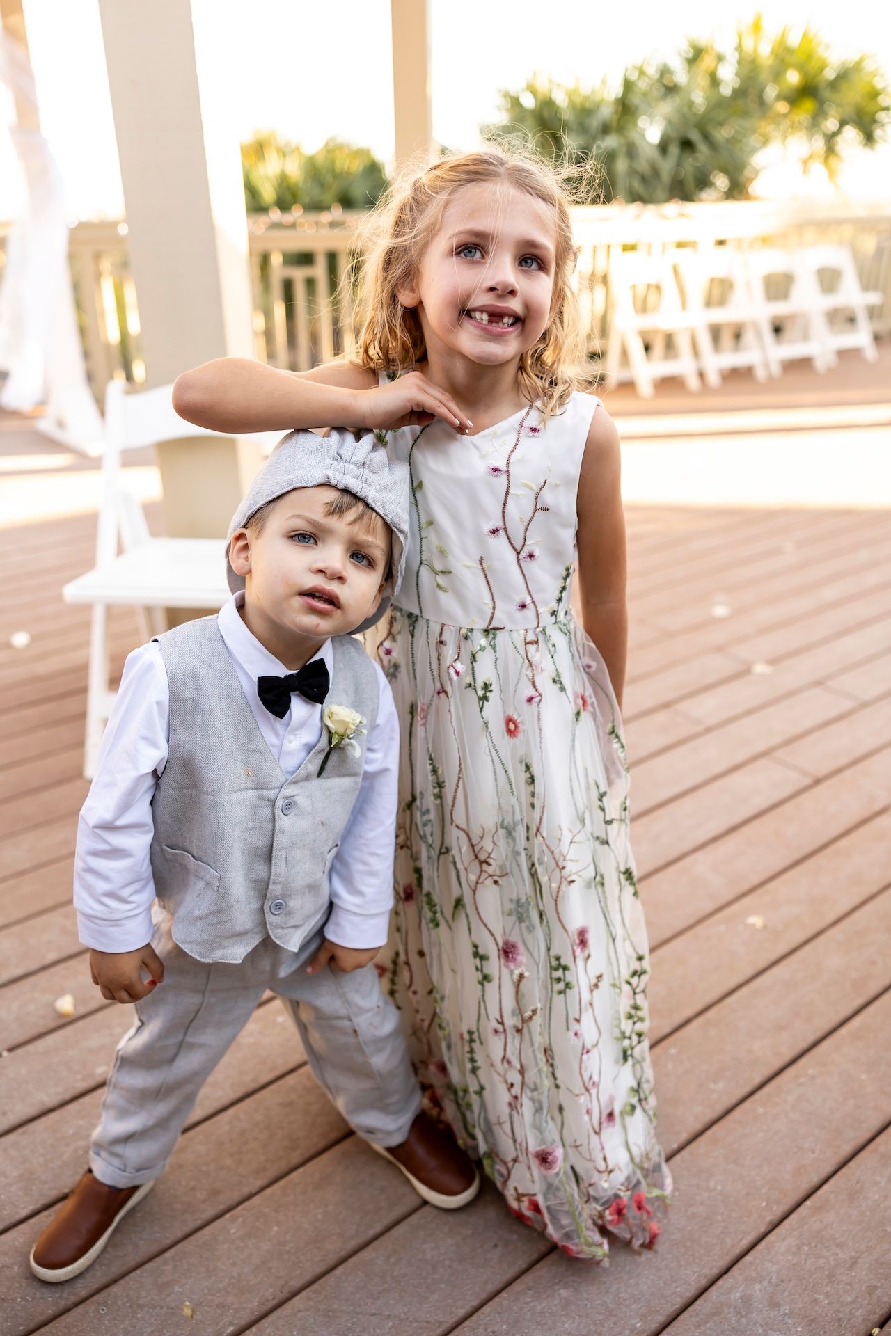 Two young children in formal attire pose on a wooden deck with palm trees visible in background.