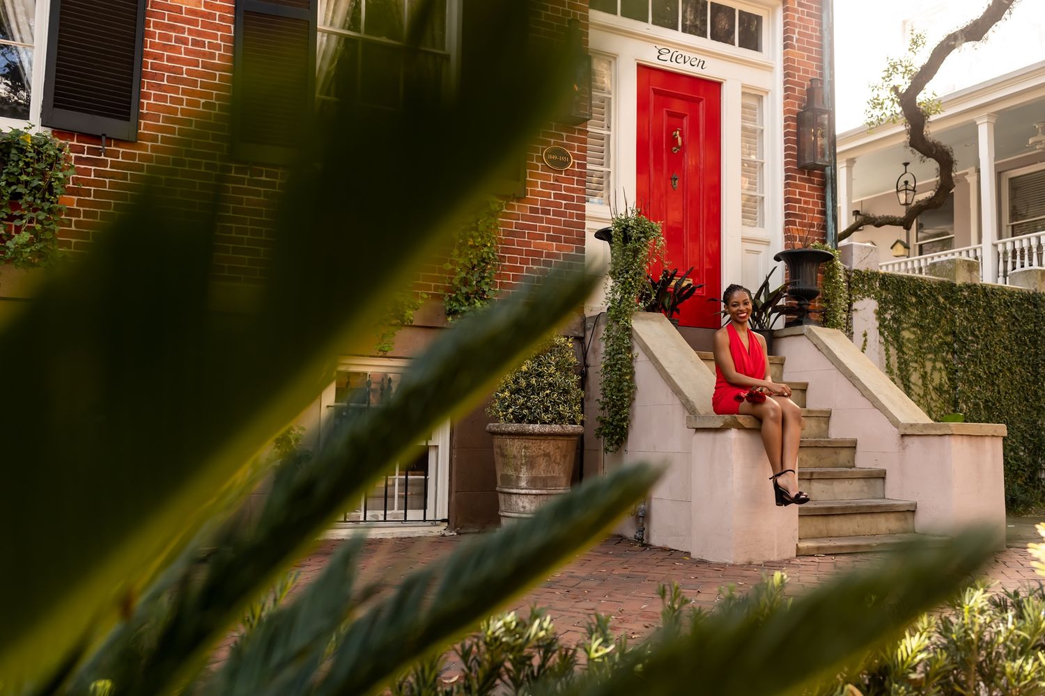 A vibrant red door adorns a brick townhouse with white steps and green foliage in the foreground.