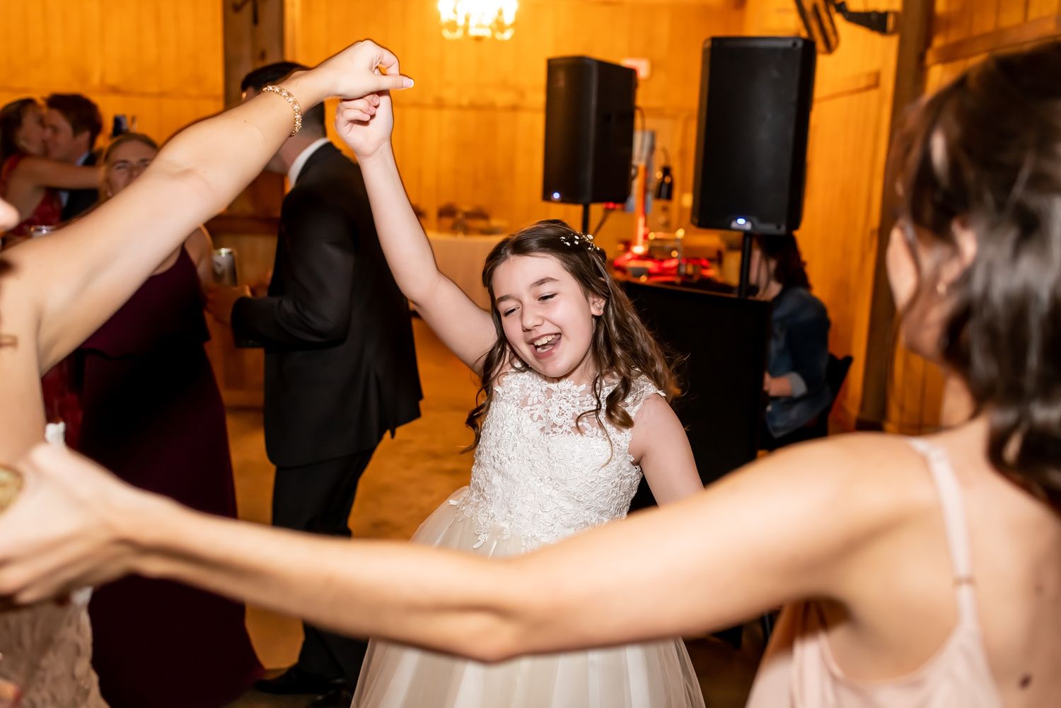 Joyful bride in white lace dress dances with guests at wedding reception in warm-lit ballroom.