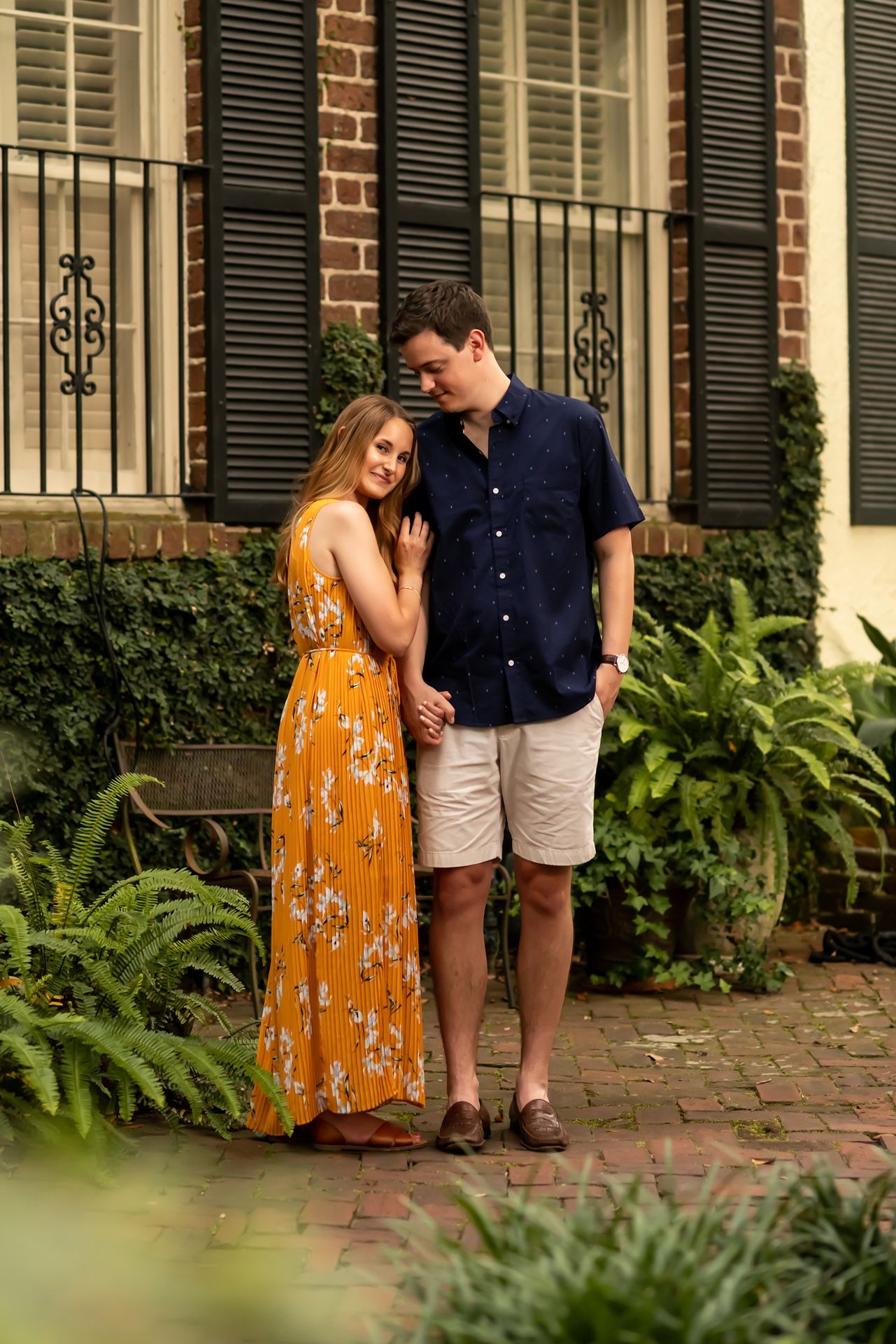 A couple in romantic pose stands on brick path by historic building with black shutters and ferns.