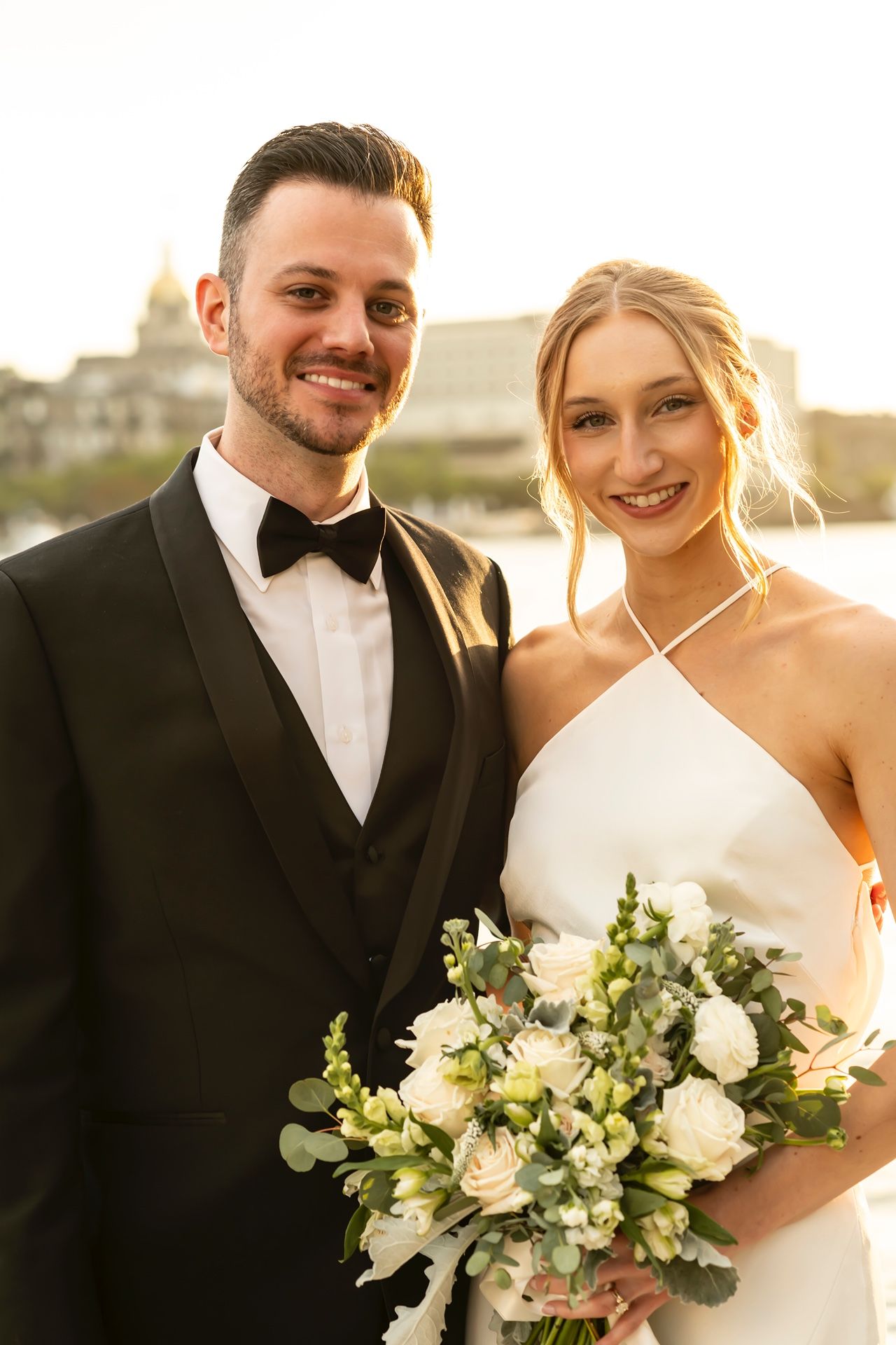 A newly married couple poses together at sunset, with the bride holding an elegant white floral bouquet.