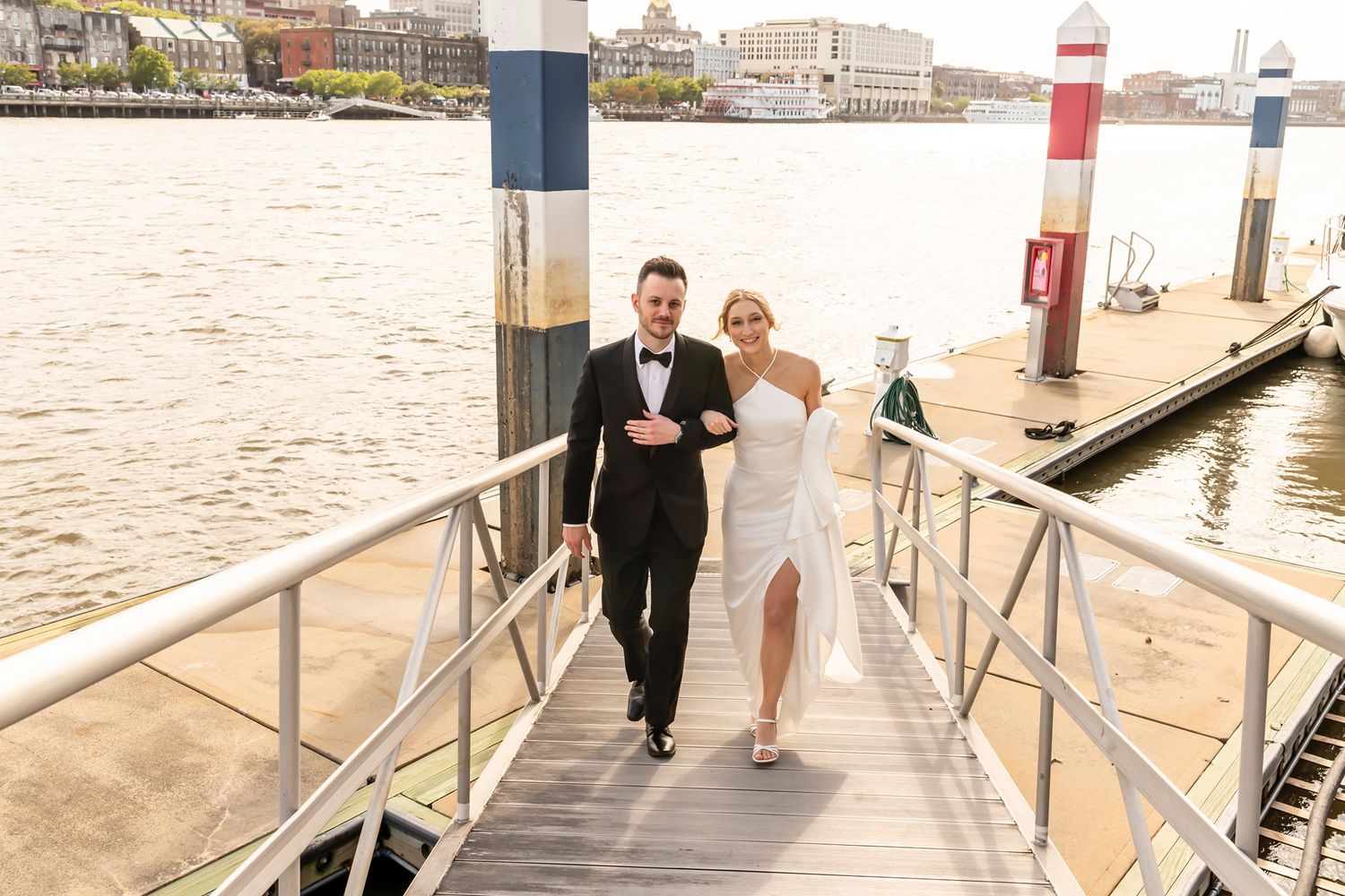 Two people walking up a boat dock ramp on a waterfront with poles and buildings in the background.