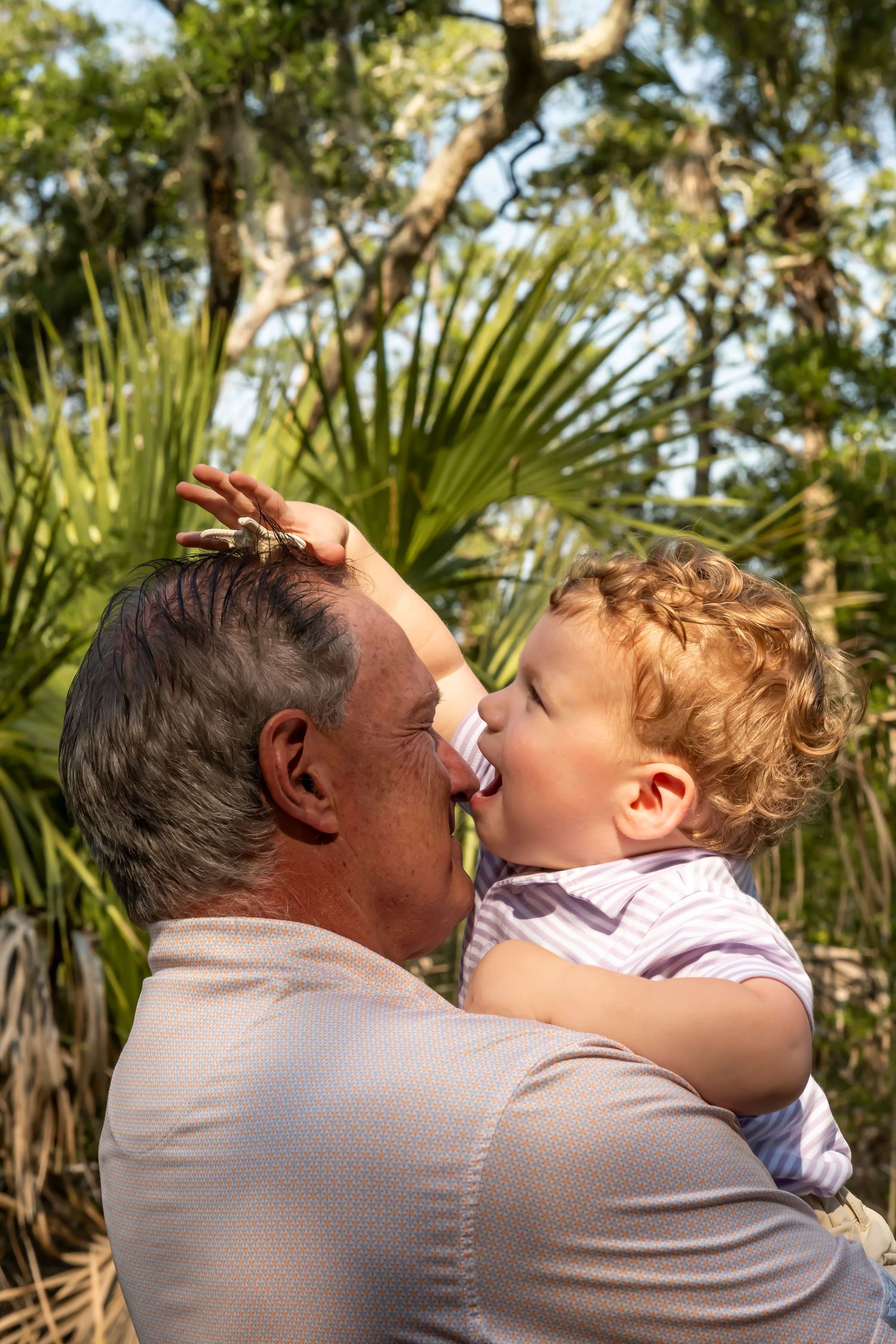 Two people share a tender moment outdoors with palm trees and greenery in the background.