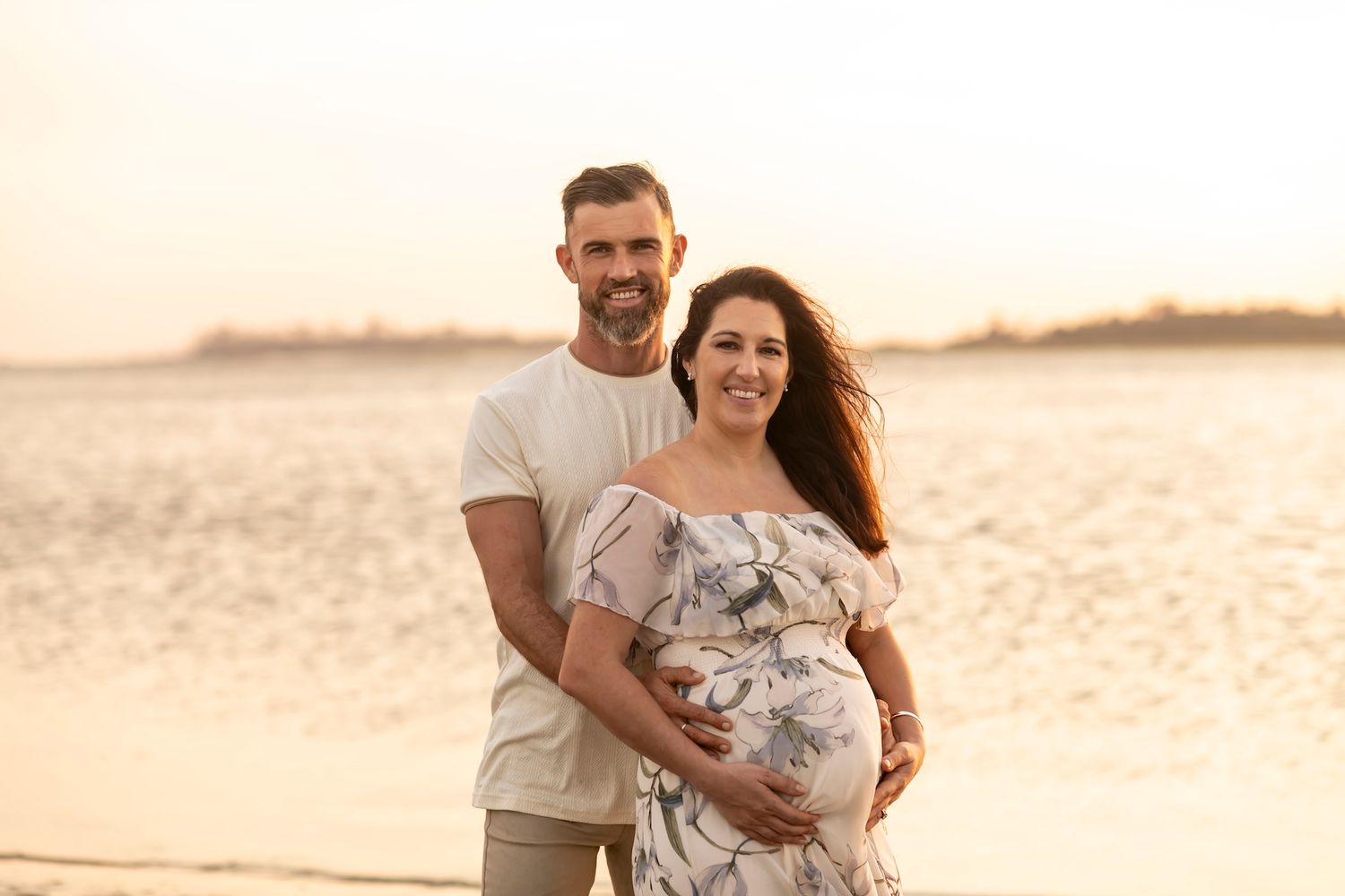 Expectant couple poses together on sandy beach during warm sunset photoshoot.