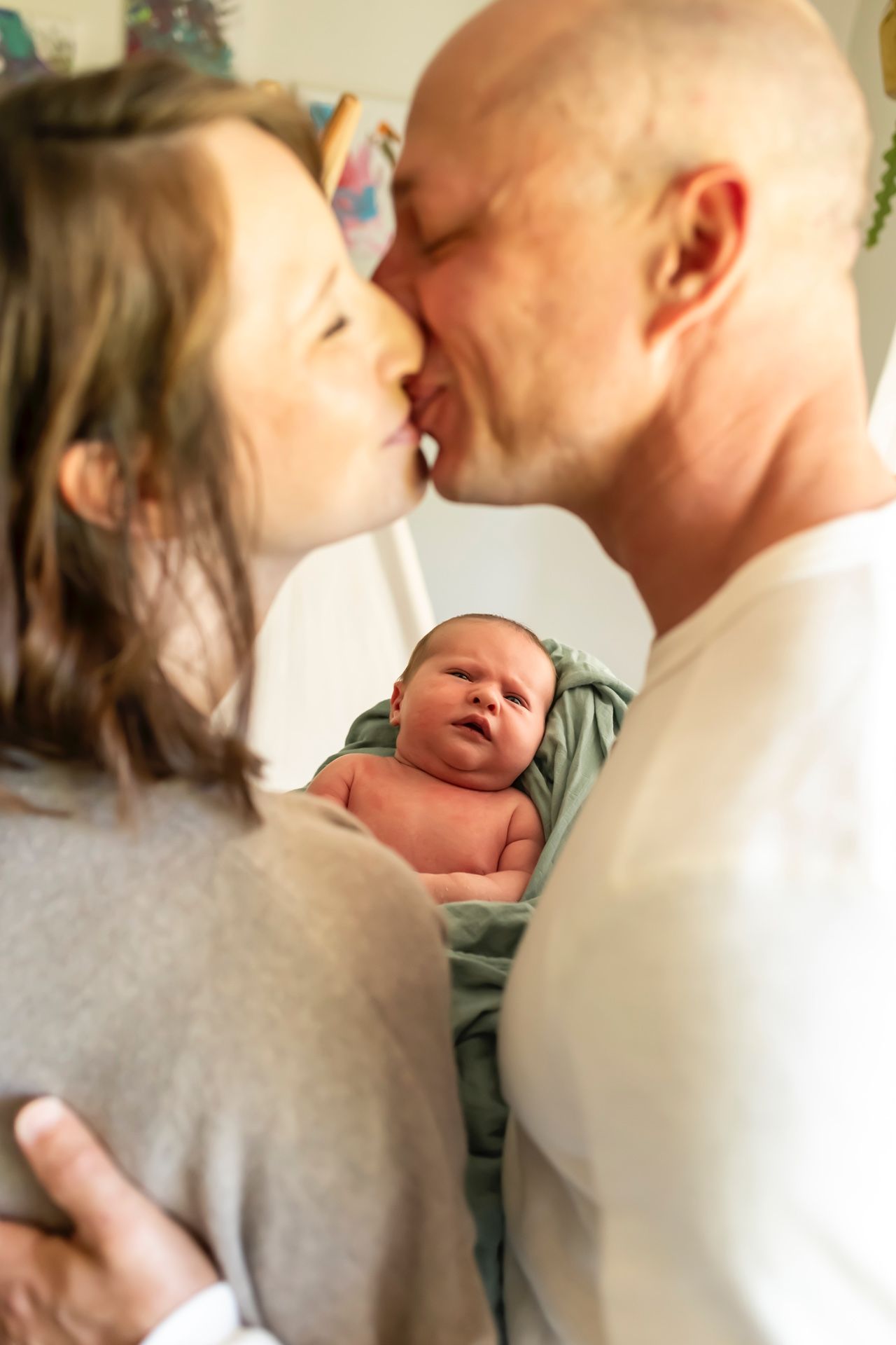 Intimate moment between parents sharing a kiss while holding their newborn baby wrapped in a mint green blanket.
