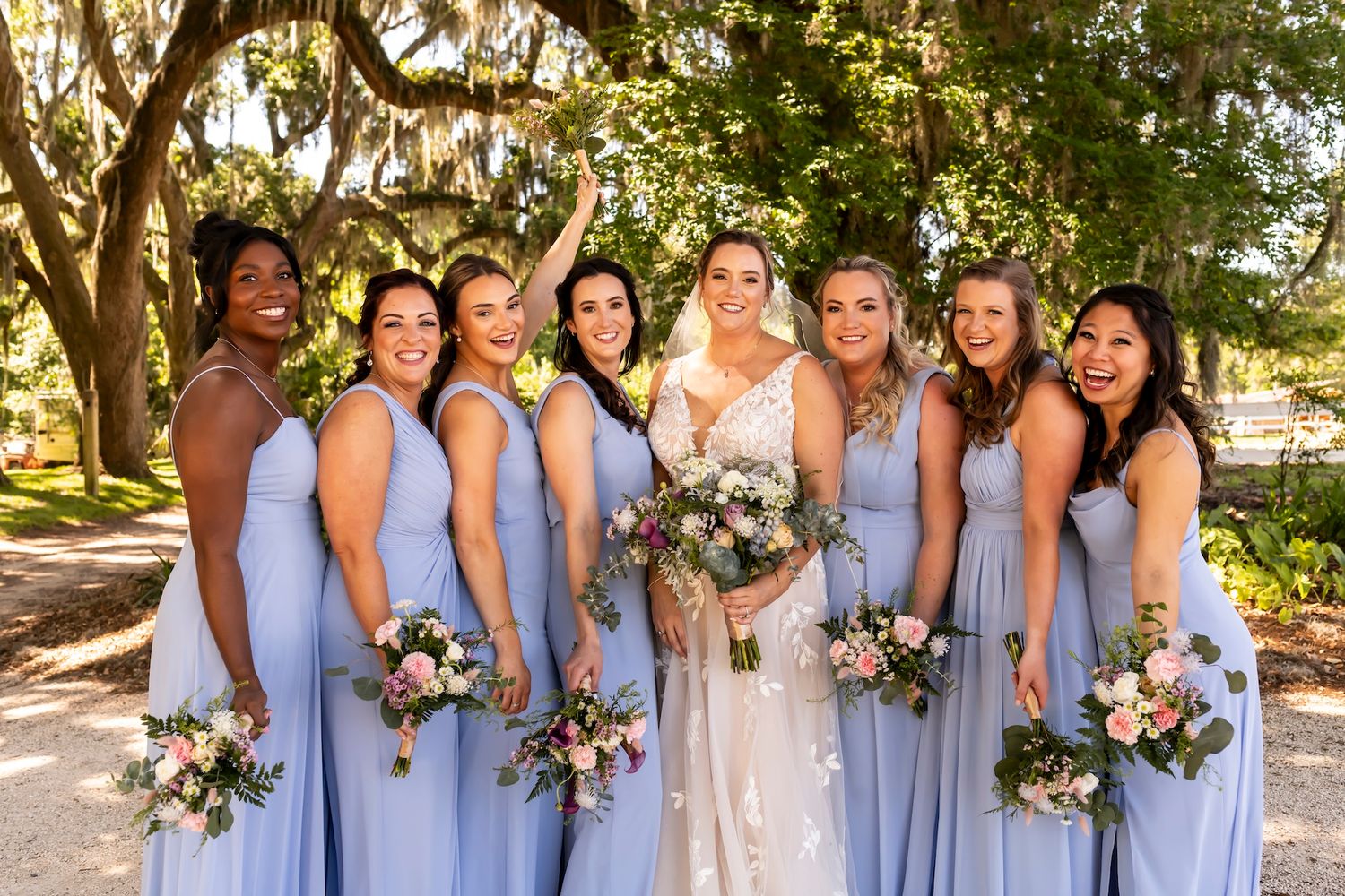 Wedding party in light blue bridesmaid dresses poses in the garden with floral bouquets.