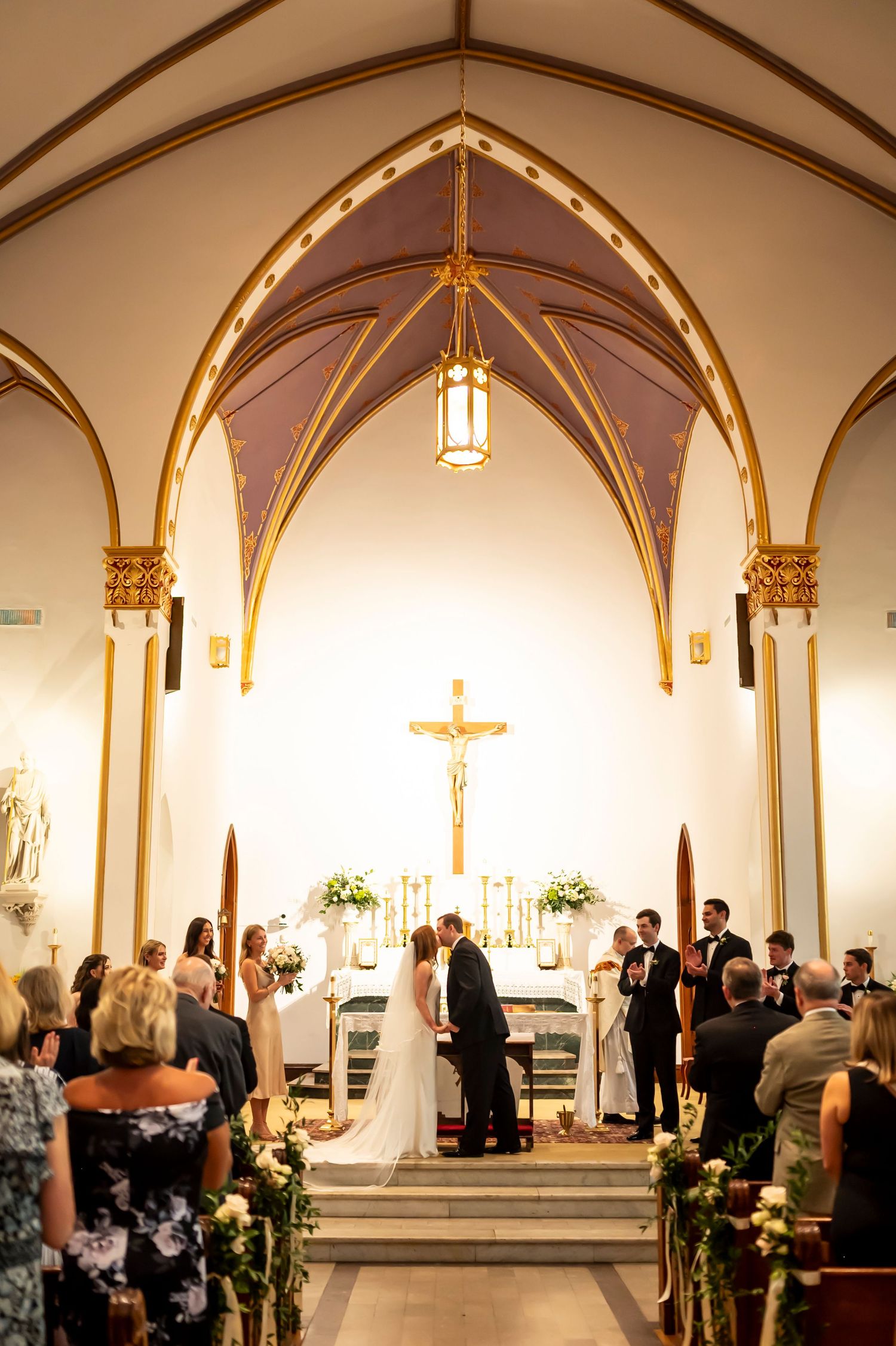 A traditional Catholic church wedding ceremony takes place under gorgeous vaulted arches with golden lighting and floral decor.