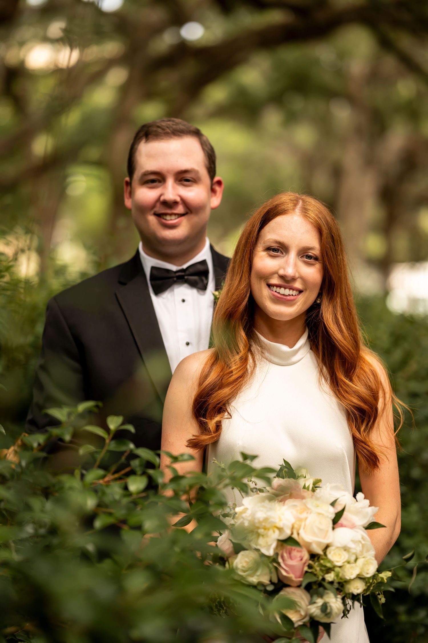 Newlywed couple in formal wedding attire stand close together in a garden setting holding a bouquet of light-colored flowers.