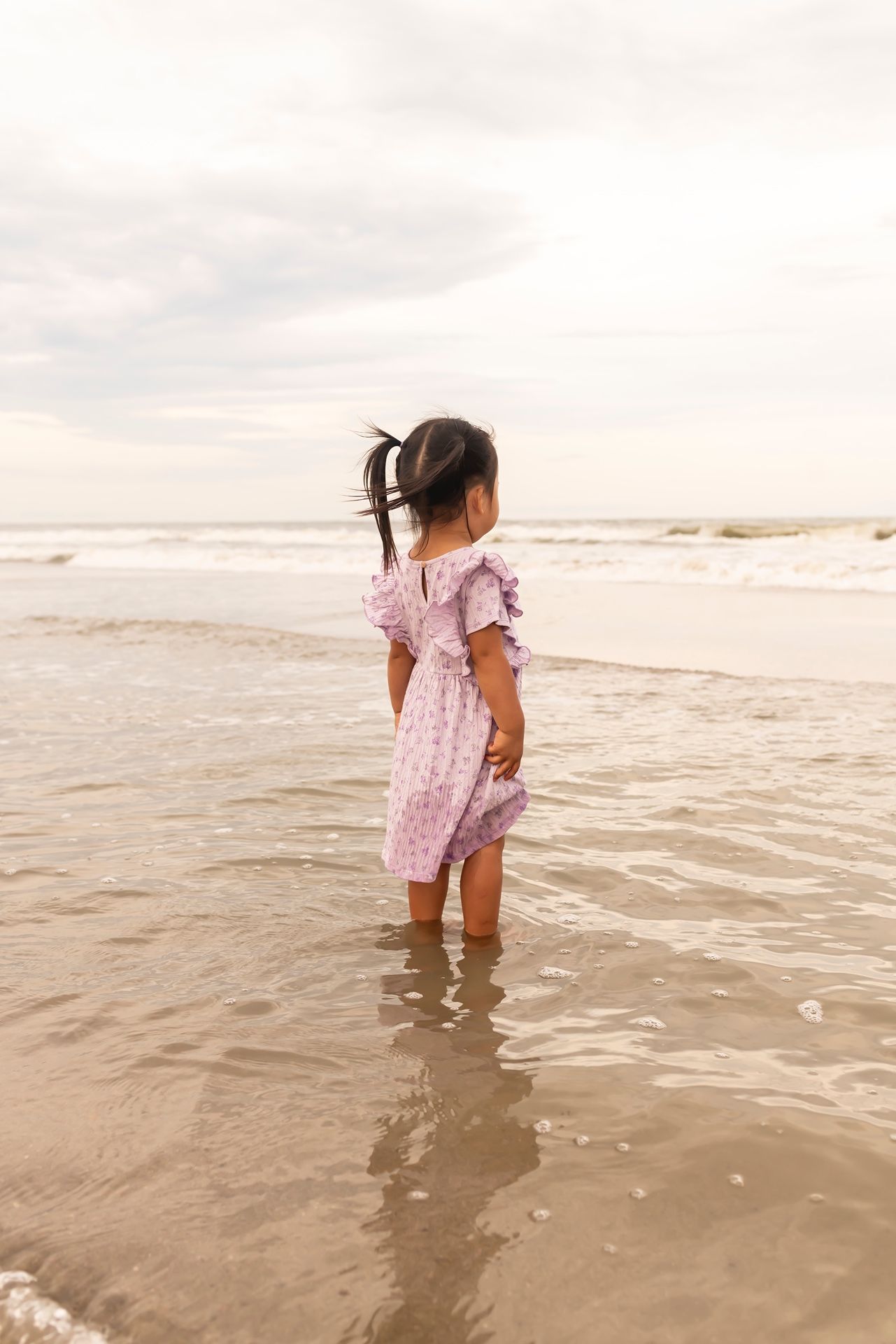 A person wearing a pink floral dress wades in shallow ocean waves at sunset.