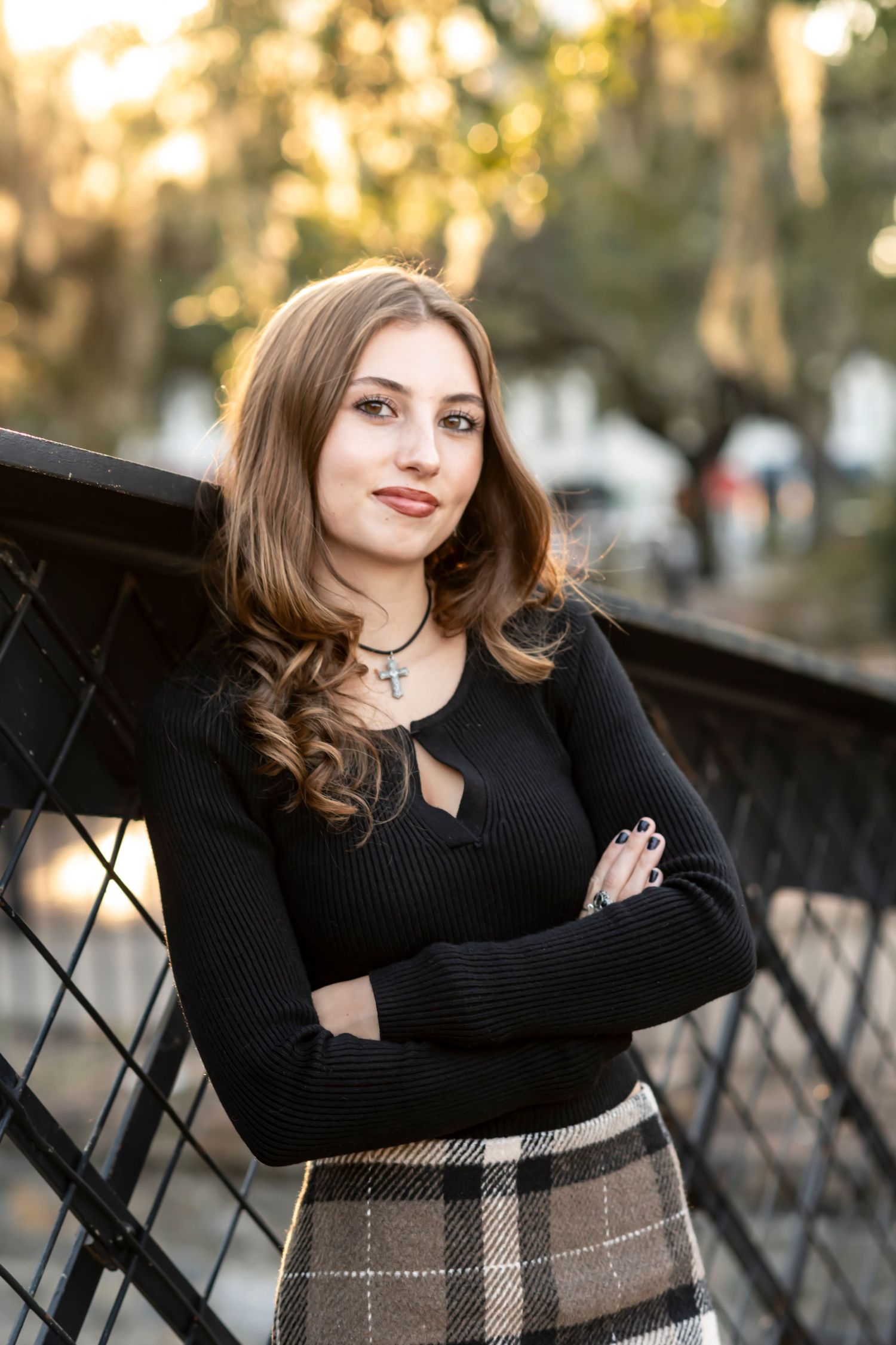 Fashionable young person in black sweater and plaid skirt poses confidently against metal railing at sunset.