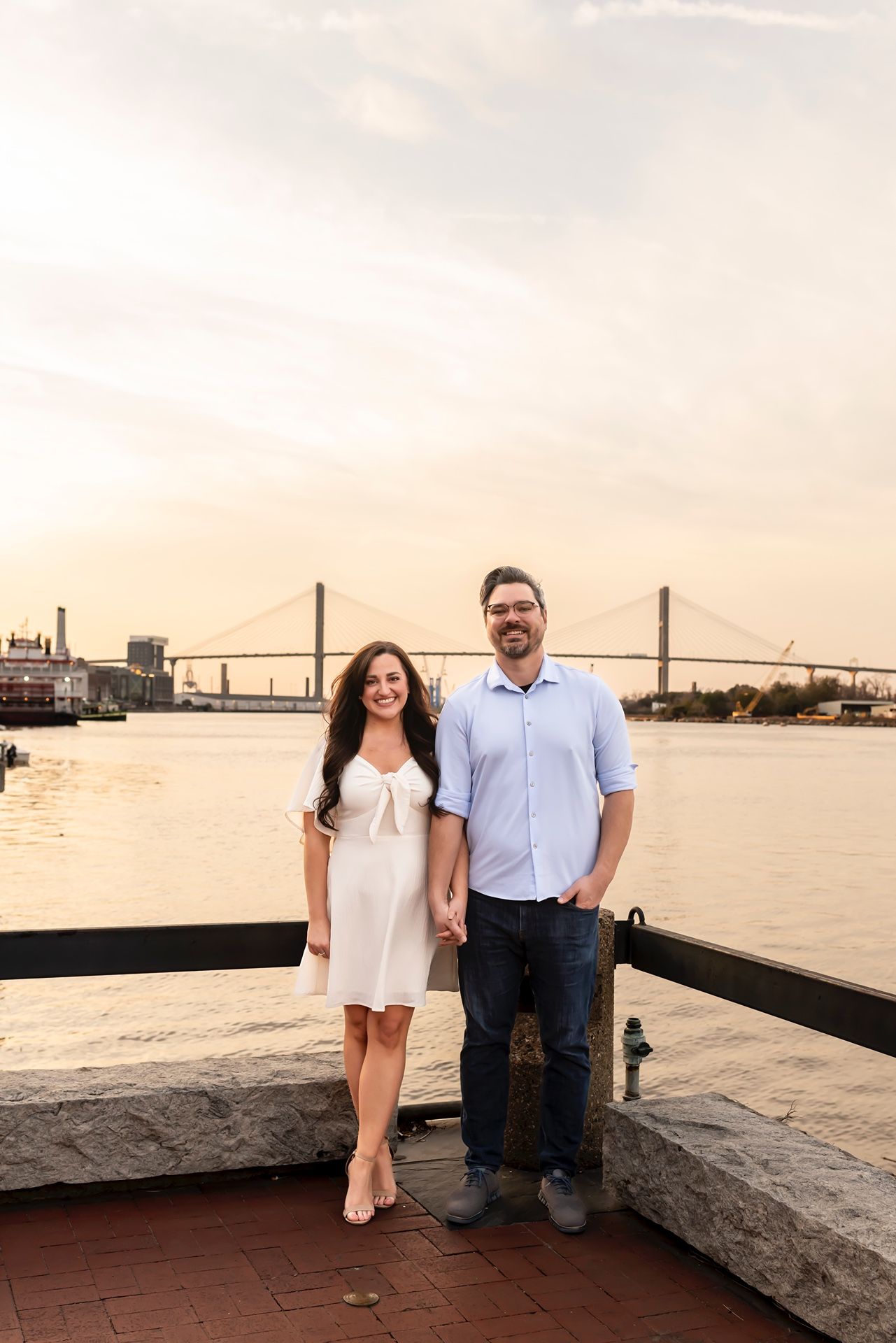 A couple stands on a brick waterfront at sunset with a large suspension bridge visible in the golden background.
