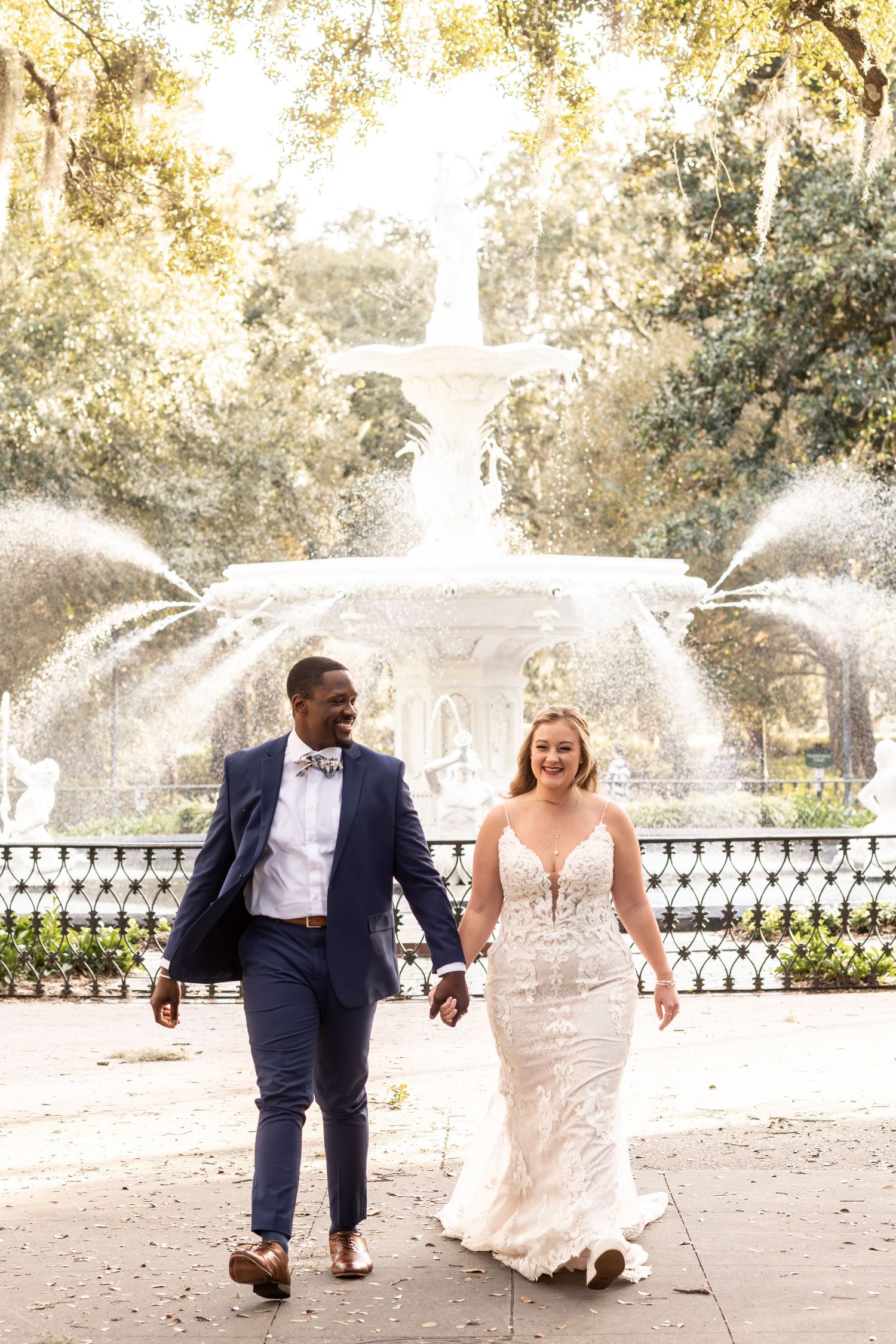 Happy newlyweds walking together in front of ornate fountain at Forsyth Park in Savannah Georgia during sunny wedding day.