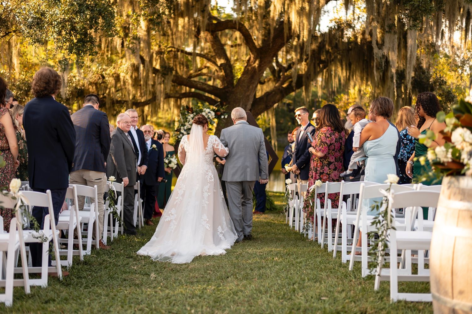 An outdoor wedding ceremony under a majestic oak tree draped in Spanish moss with white chairs along the aisle at sunset.