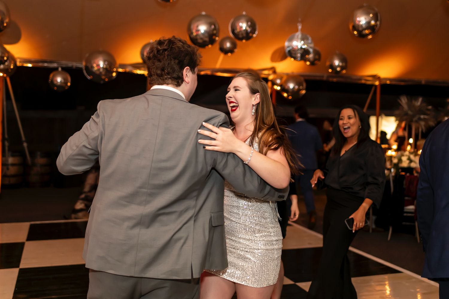 Two people dance together on a checkered floor under disco balls at a lively party celebration.