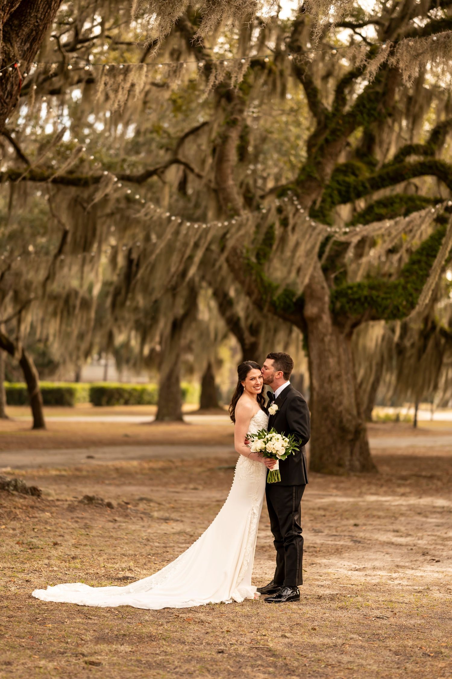 A romantic couple stands beneath Spanish moss-draped oak trees in a scenic outdoor setting with string lights.