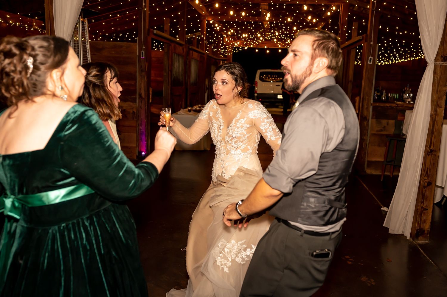 People dance together at a wedding reception under romantic string lights in a rustic barn venue.