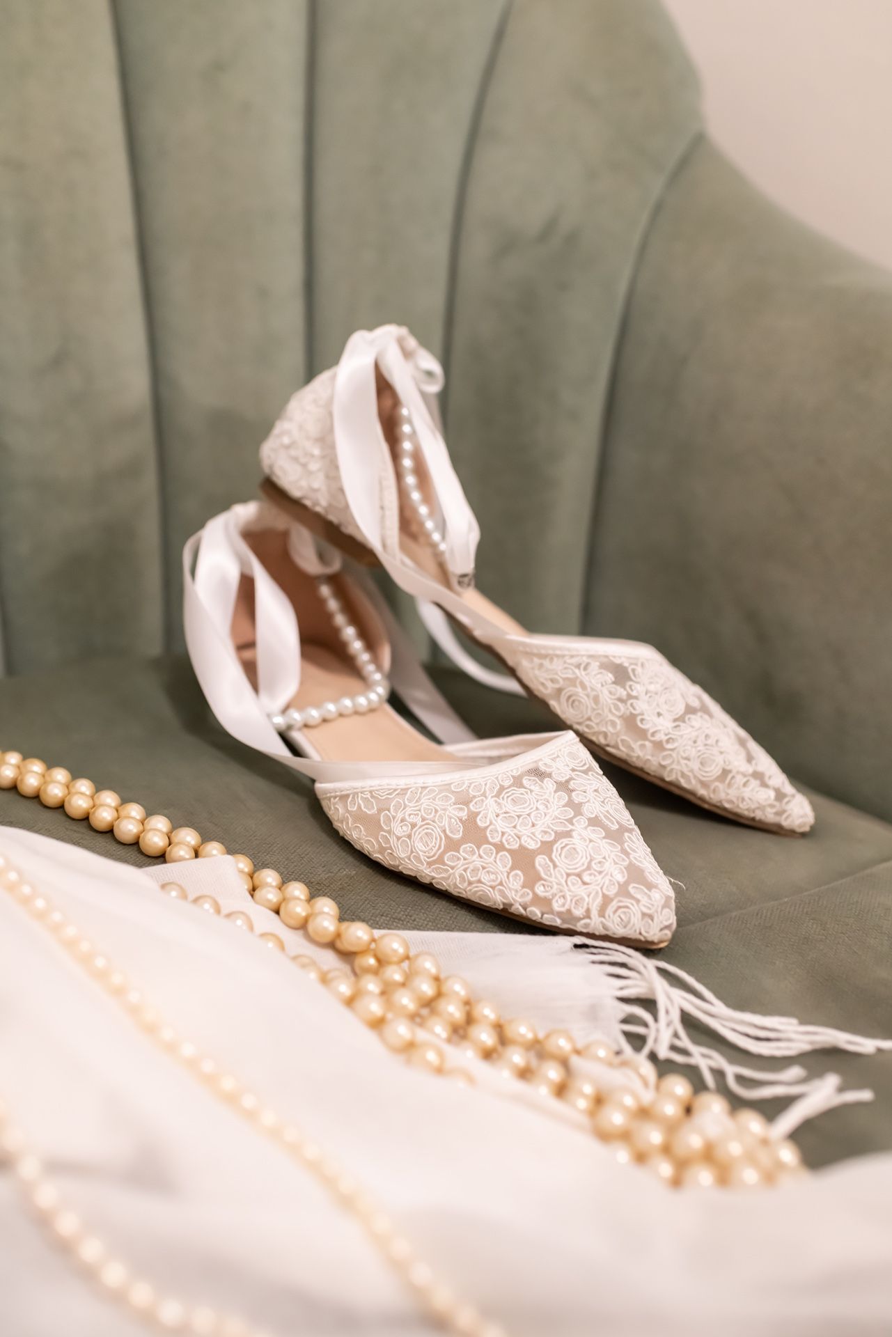A string of pearls necklace rests beside elegant blush pink lace bridal flats on a grey velvet surface.
