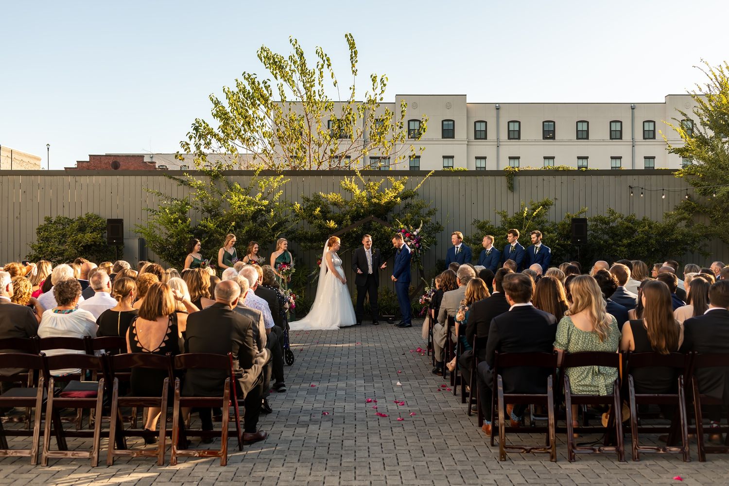 An outdoor wedding ceremony at sunset with guests seated in rows on a stone patio and buildings in the background.