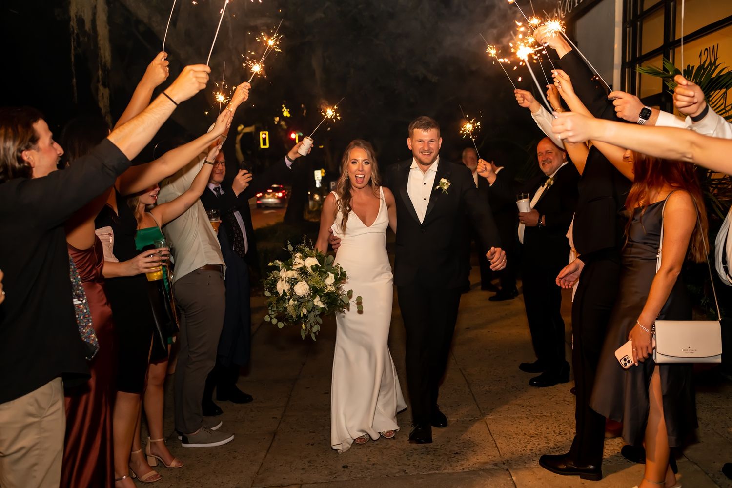 Newlyweds walk through sparkler exit line at nighttime wedding celebration with guests lining pathway.