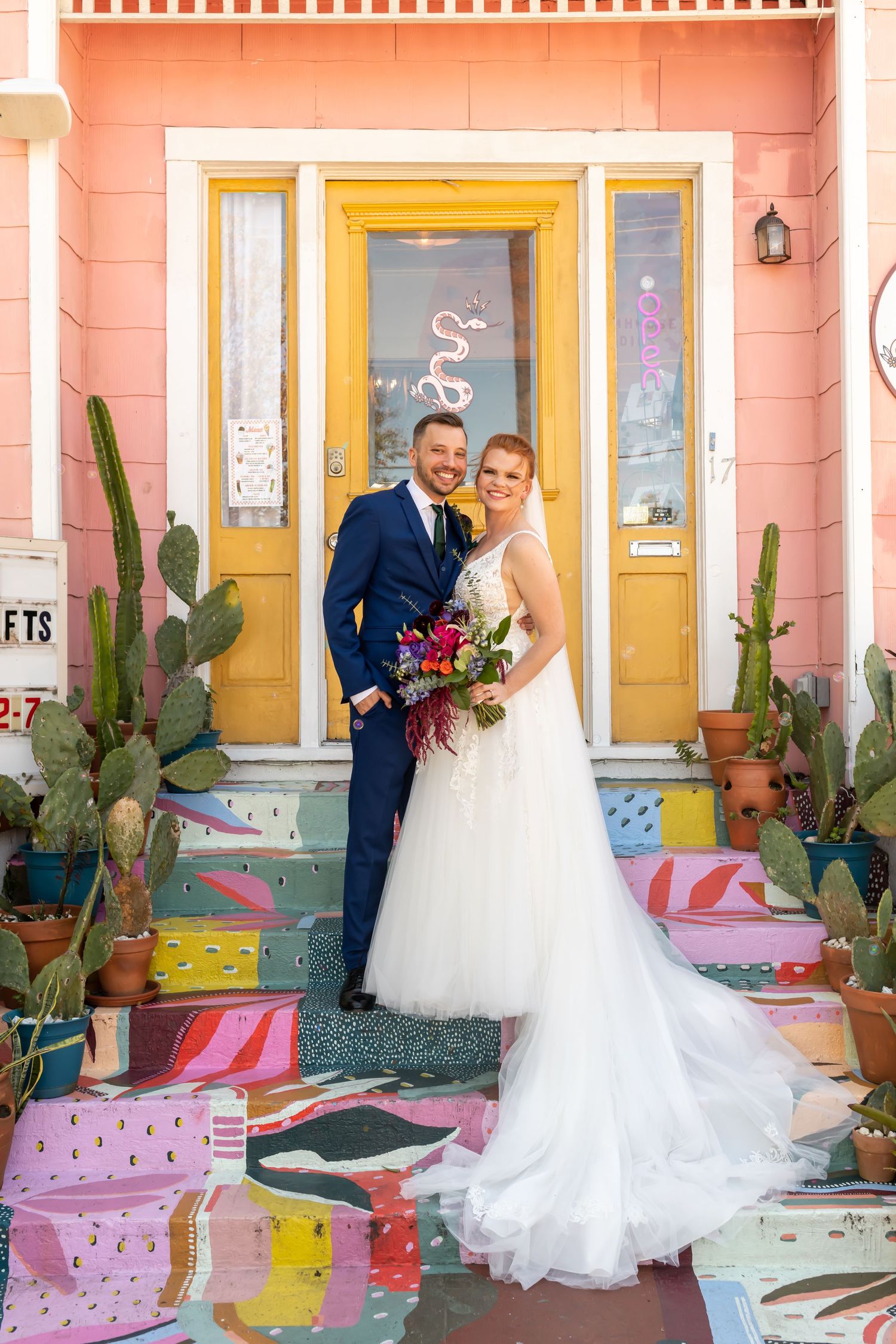 Wedding couple stands on colorful painted steps in front of pink house with yellow door and potted cacti.