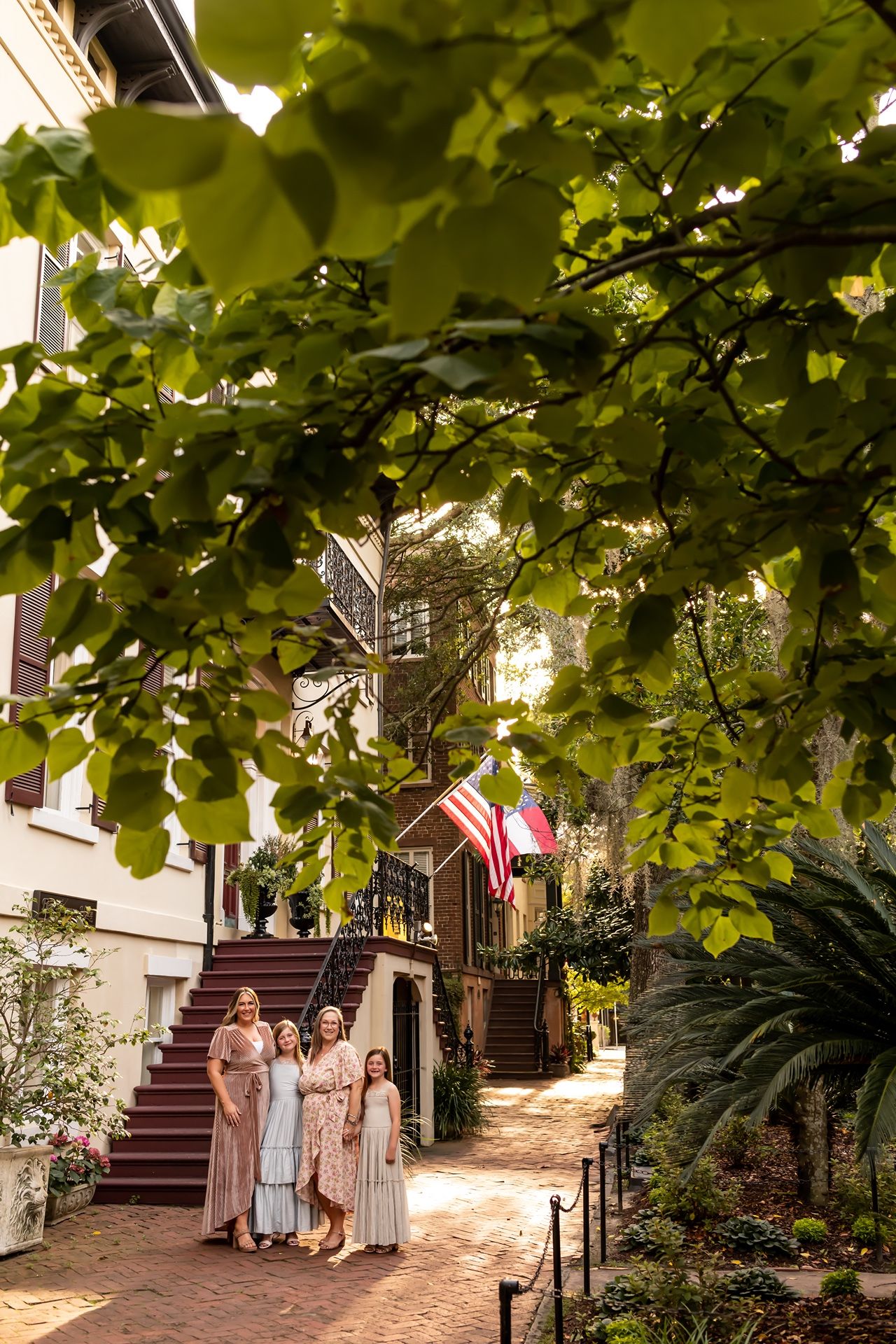 Group of people standing on a brick path under lush green trees at sunset near historic building steps.