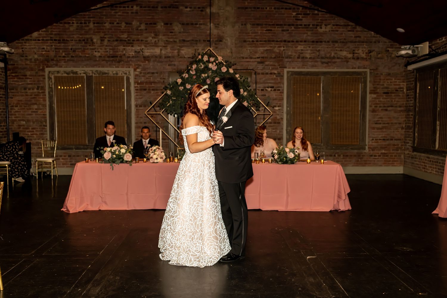 A couple shares their first dance at a rustic indoor reception venue with pink tablecloths and holiday decor in background.
