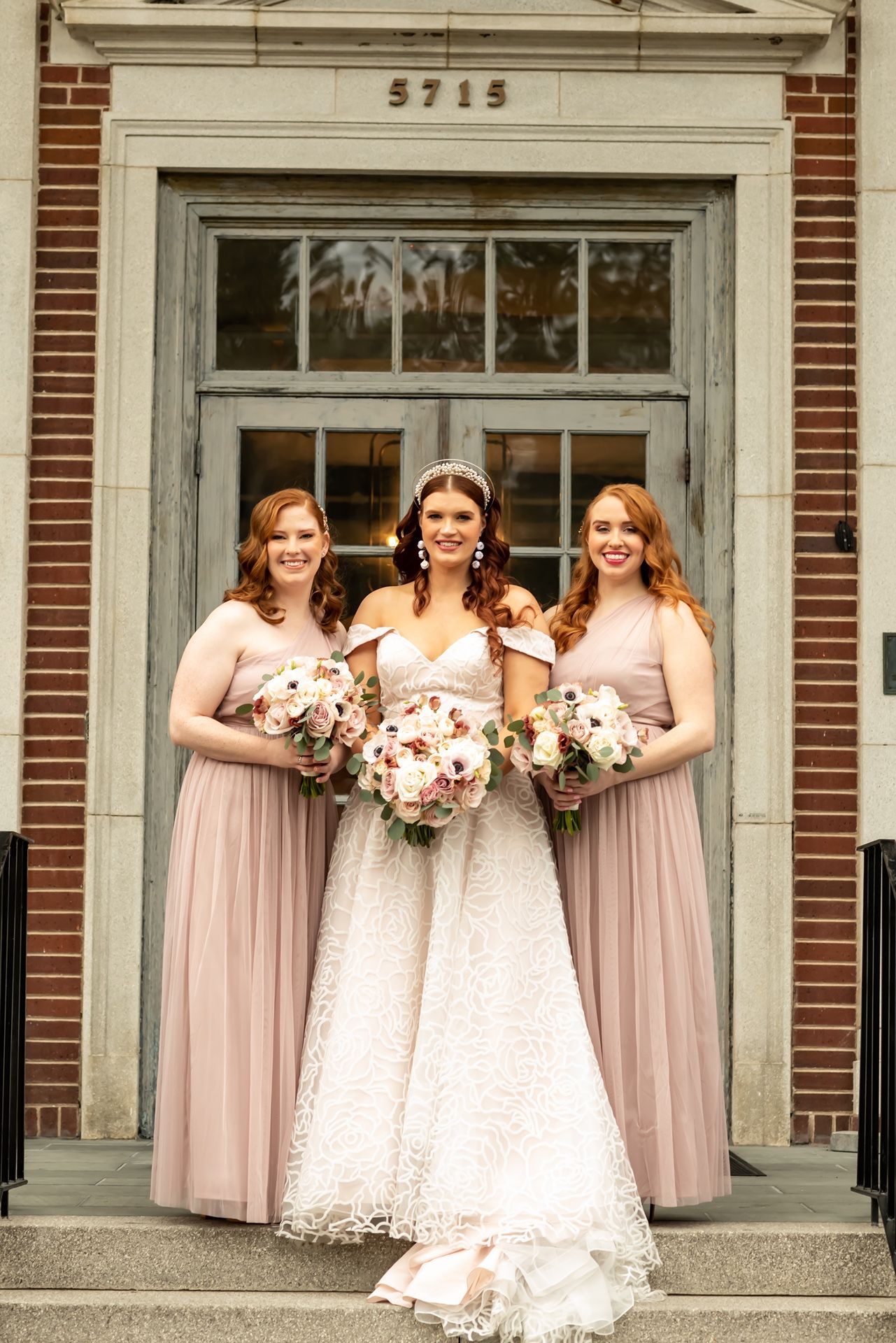 Bridesmaids wearing pink dresses and holding floral bouquets pose together on brick building steps.