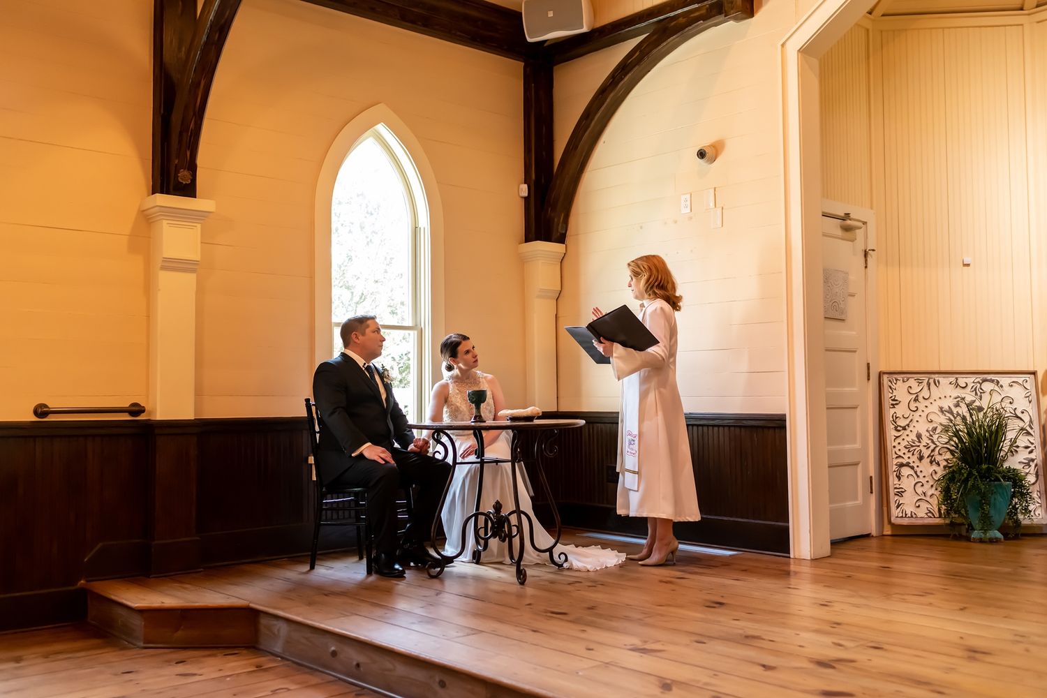 Three people perform music together in a bright church-like hall with wooden floors and arched windows.