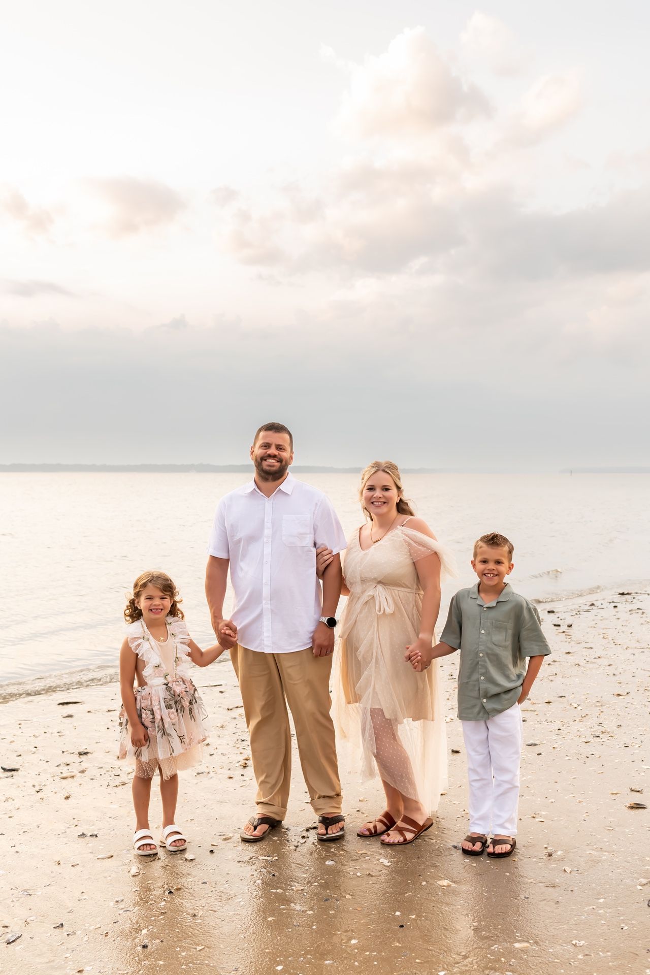 A family of five stands together on a serene beach at sunset wearing coordinated neutral-colored outfits.