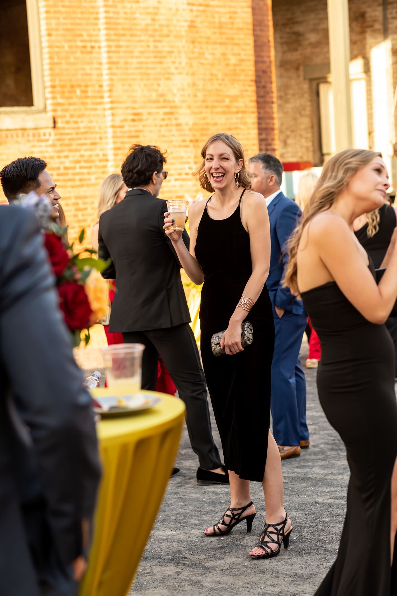A group of elegantly dressed people socializing at an event with brick walls and yellow accents in the background.