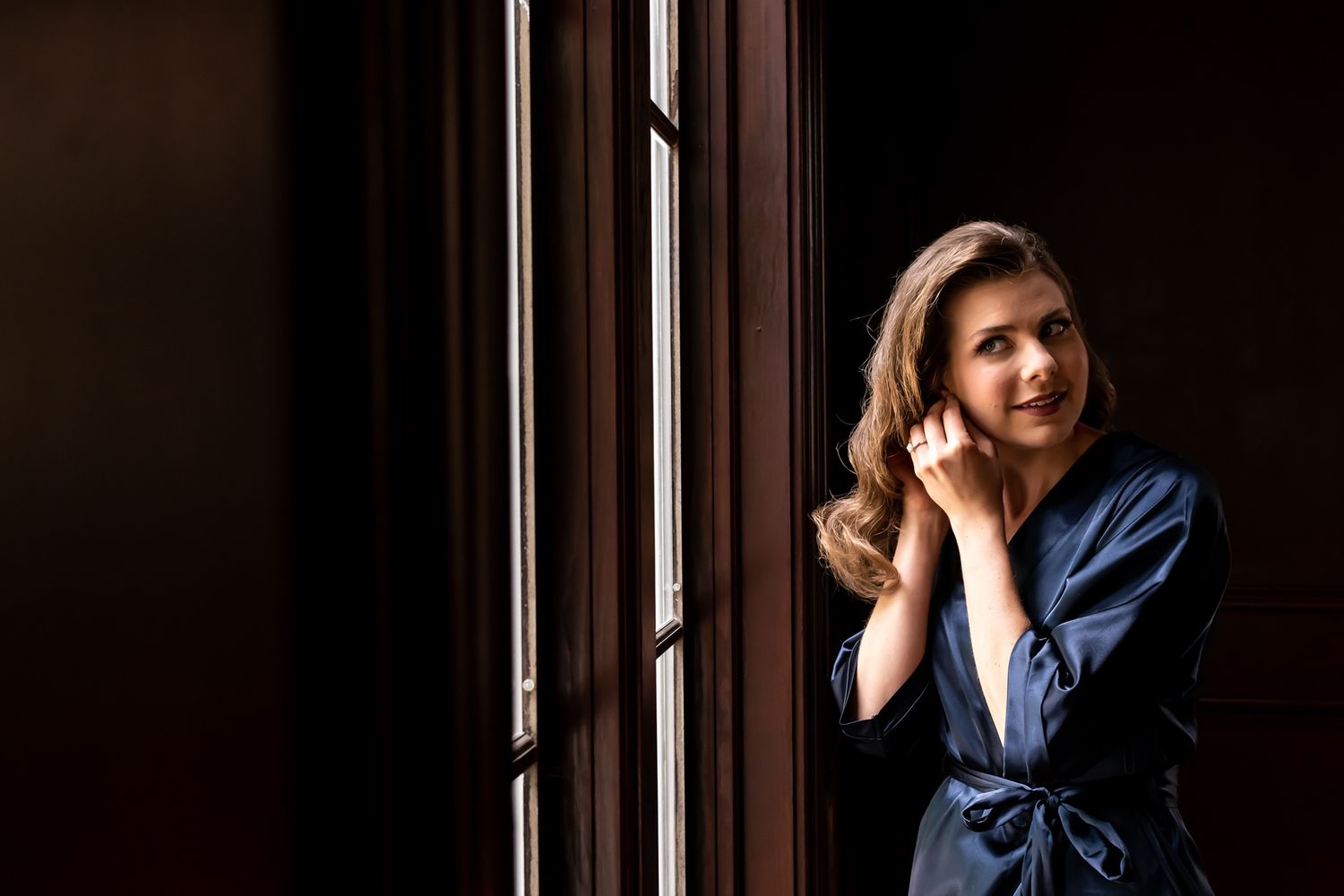 A person in a blue robe stands by a window in dramatic lighting while touching their hair.