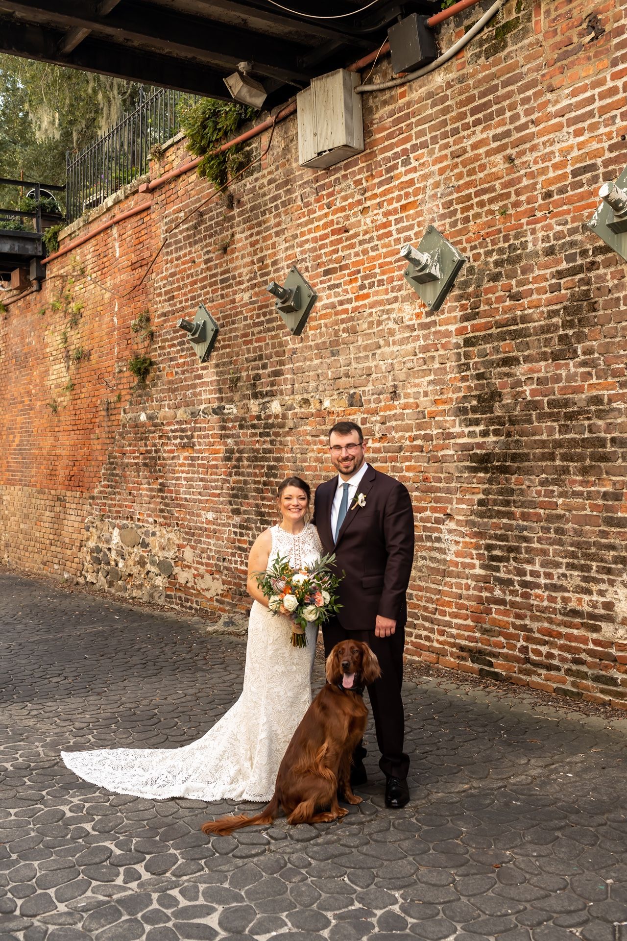 A couple poses with their brown dog in formal attire against a rustic brick wall for their wedding portrait.