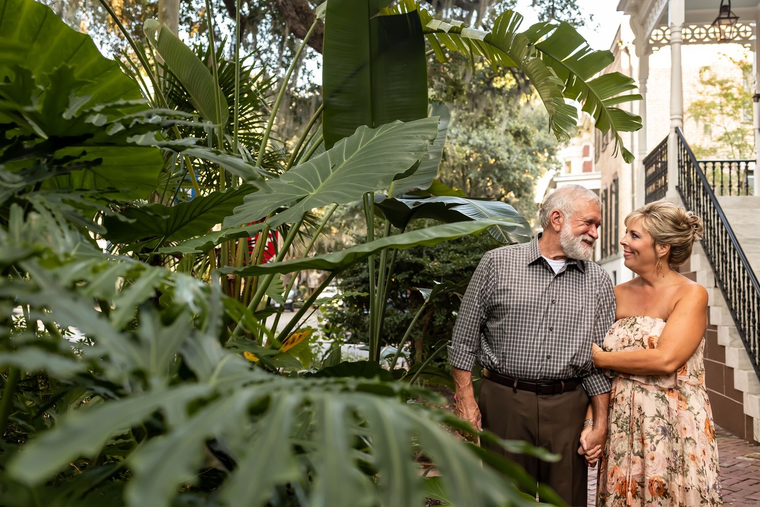 Two people share an affectionate moment together while standing among tropical plants in a garden setting.