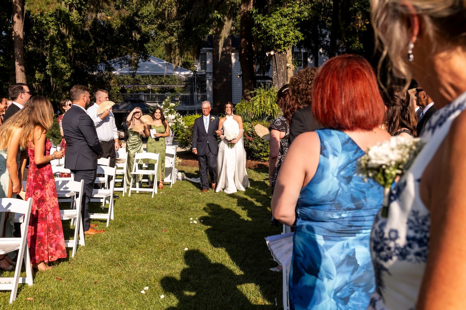 Outdoor wedding ceremony with guests seated in white chairs as bride walks down the aisle on a sunny day.