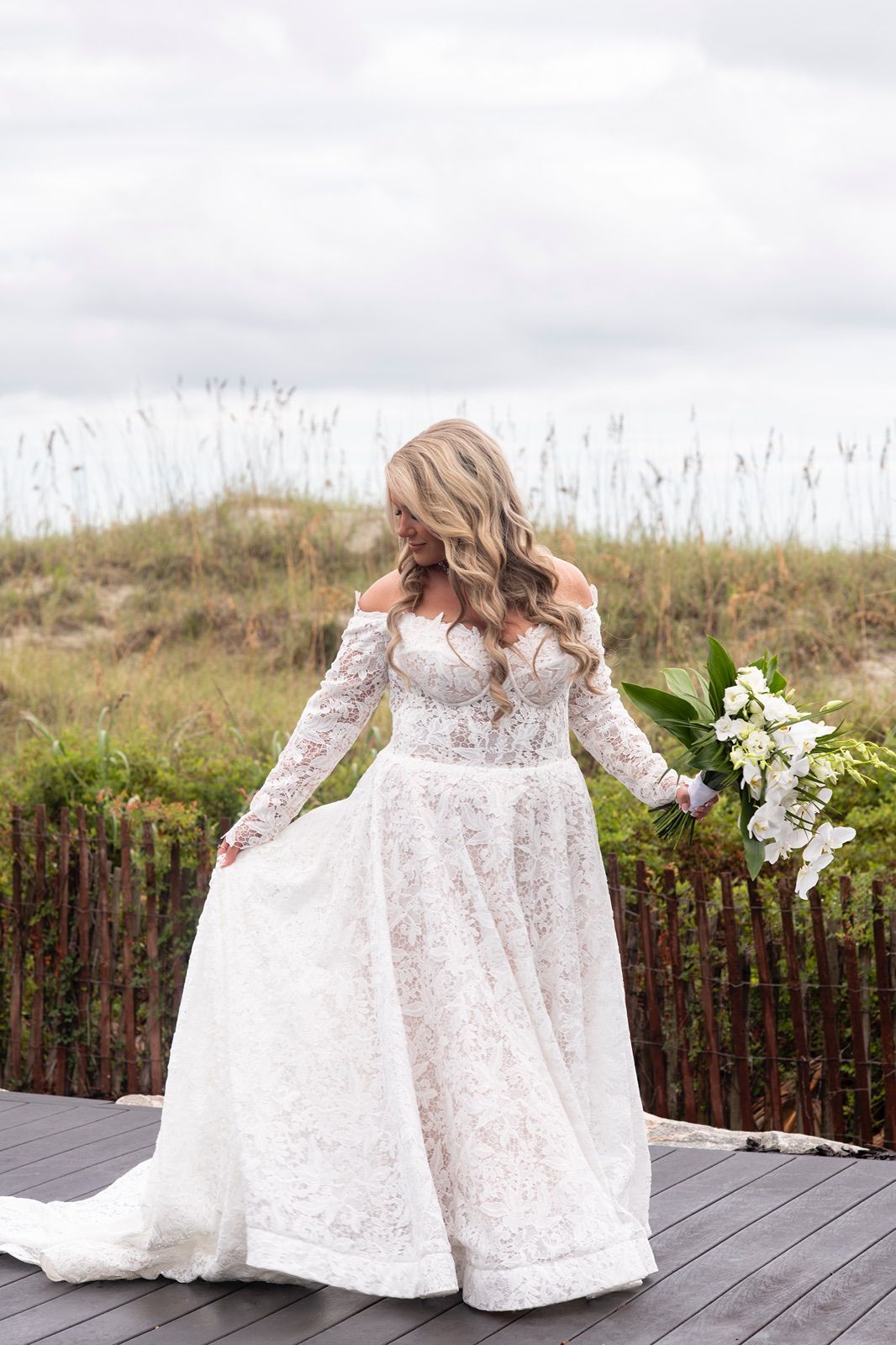 White lace wedding dress with long sleeves flowing in the wind on a wooden deck with scenic background.
