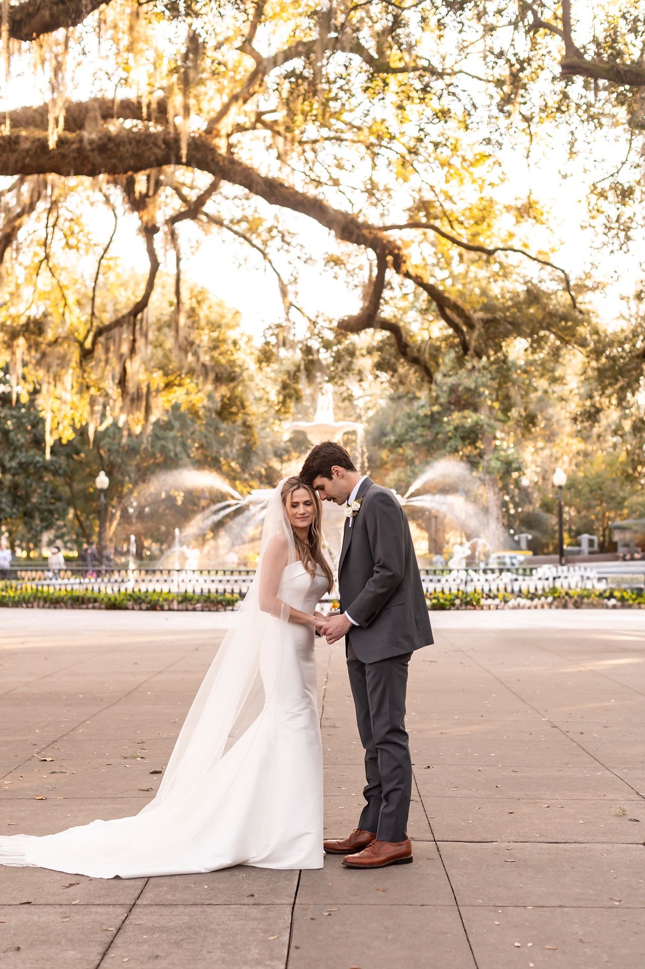 Romantic wedding couple embraces during golden hour sunset under moss-draped oak trees in a Southern garden.