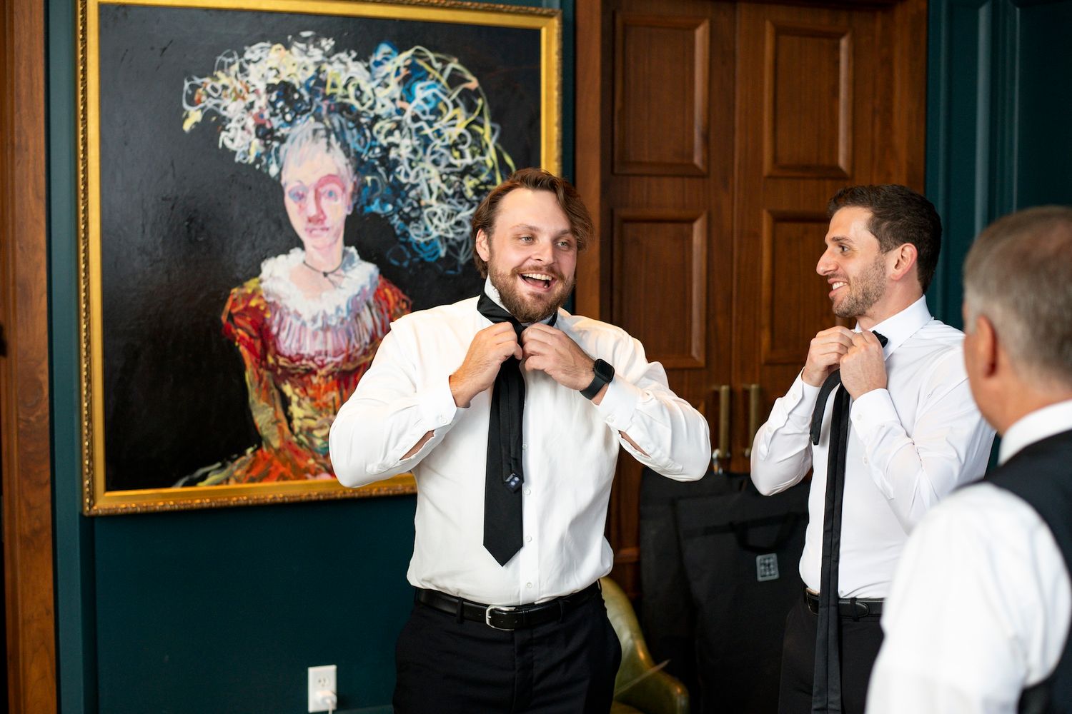 A group of people in formal attire laugh and interact during what appears to be a wedding reception or formal event.