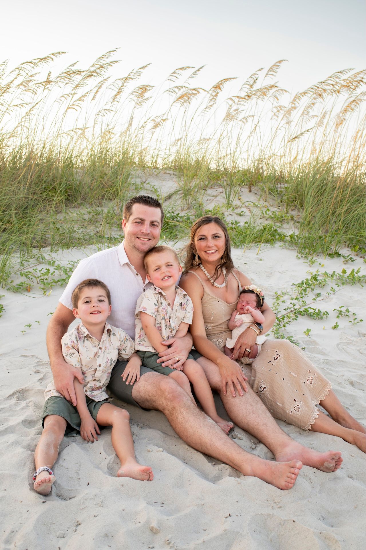 Family of five poses on sandy beach with sea oats, parents with two young boys and newborn baby, casual coastal portrait.