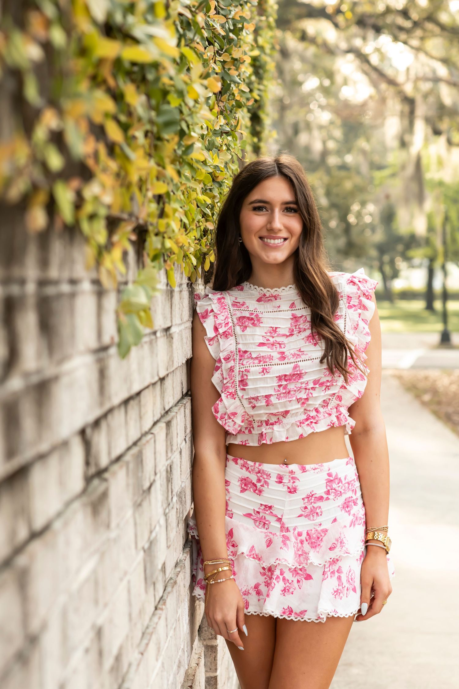Woman wearing matching pink floral crop top and skirt set standing against stone wall.