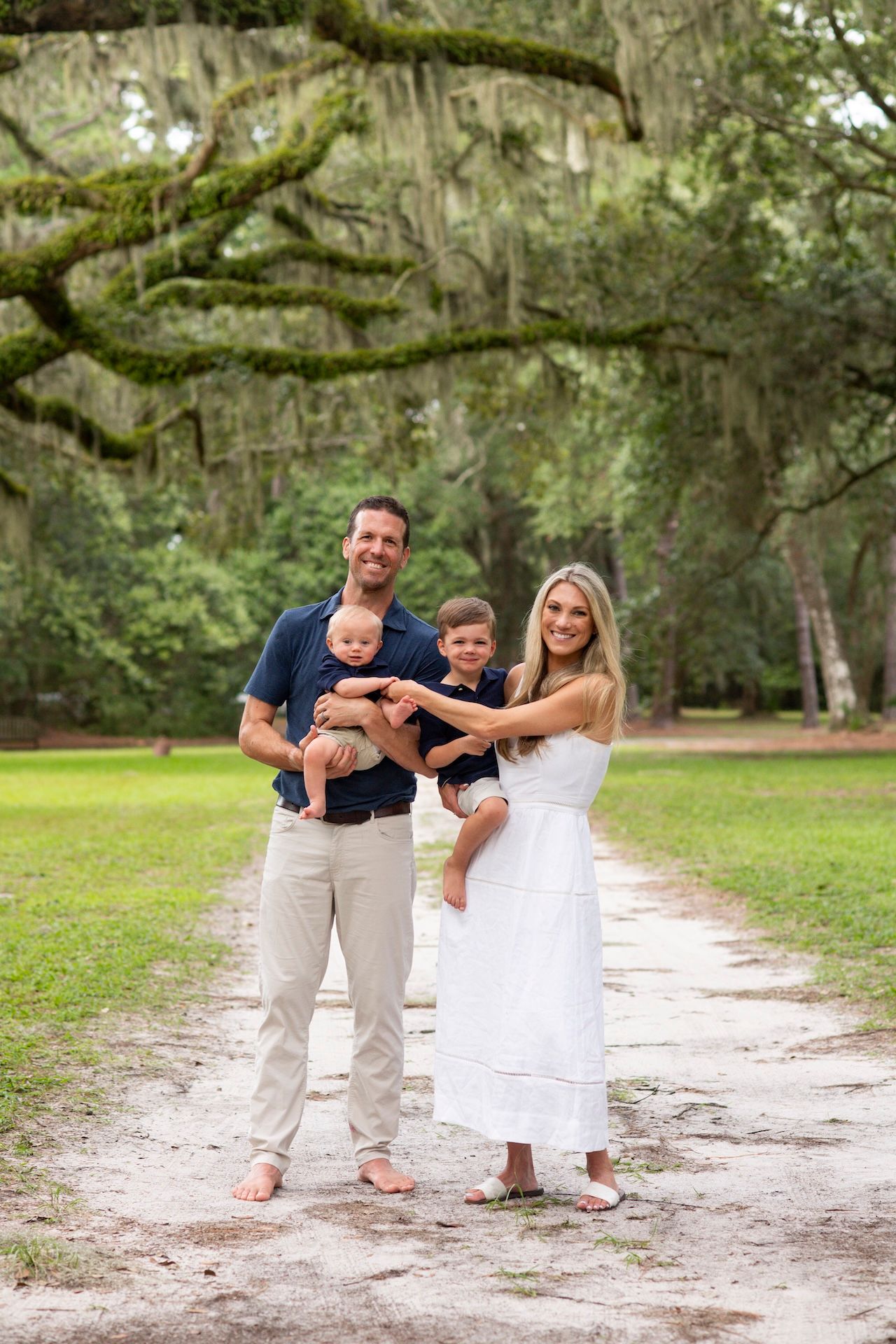 Family of four posing on a tree-lined path with Spanish moss, parents holding two young children outdoors.