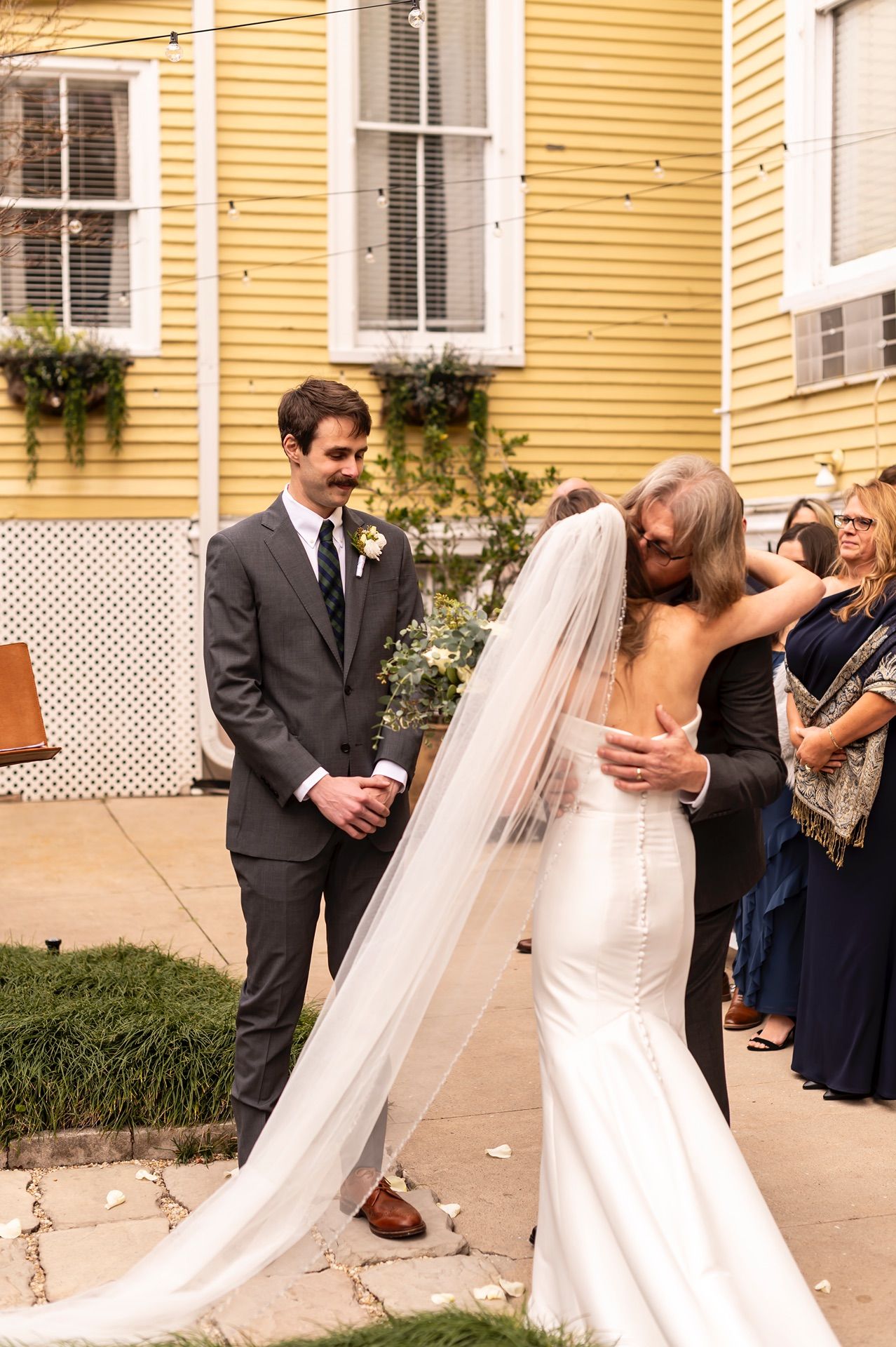 A wedding celebration takes place in front of a charming yellow historic house with white shutters.