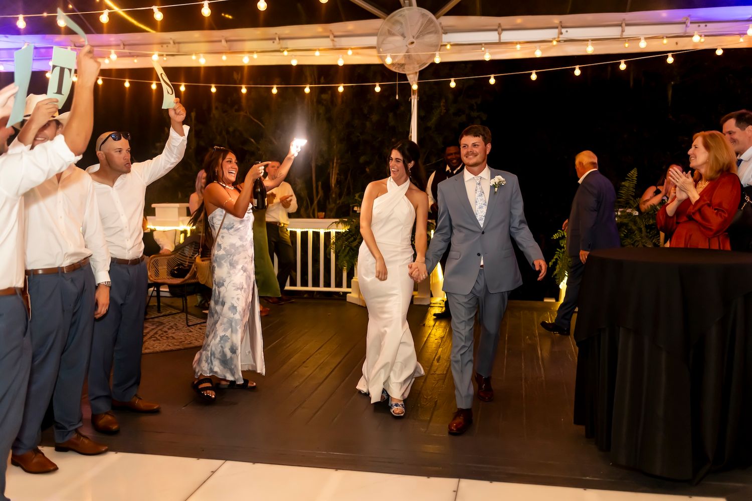 Wedding guests cheer and celebrate on a dance floor under twinkling lights at an evening reception.
