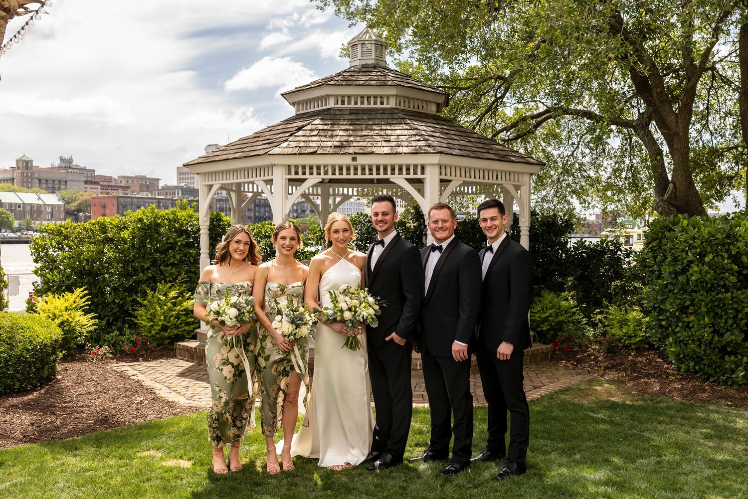 Wedding party poses together on a lawn in front of an elegant octagonal gazebo on a sunny day.