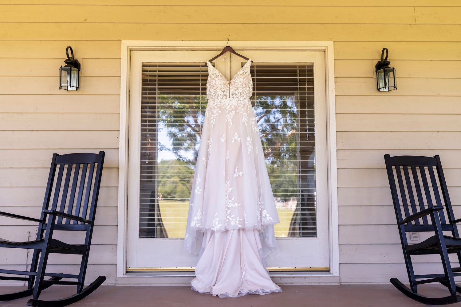 A rustic front porch with black rocking chairs and a wedding dress hanging at a Southern home with yellow siding.