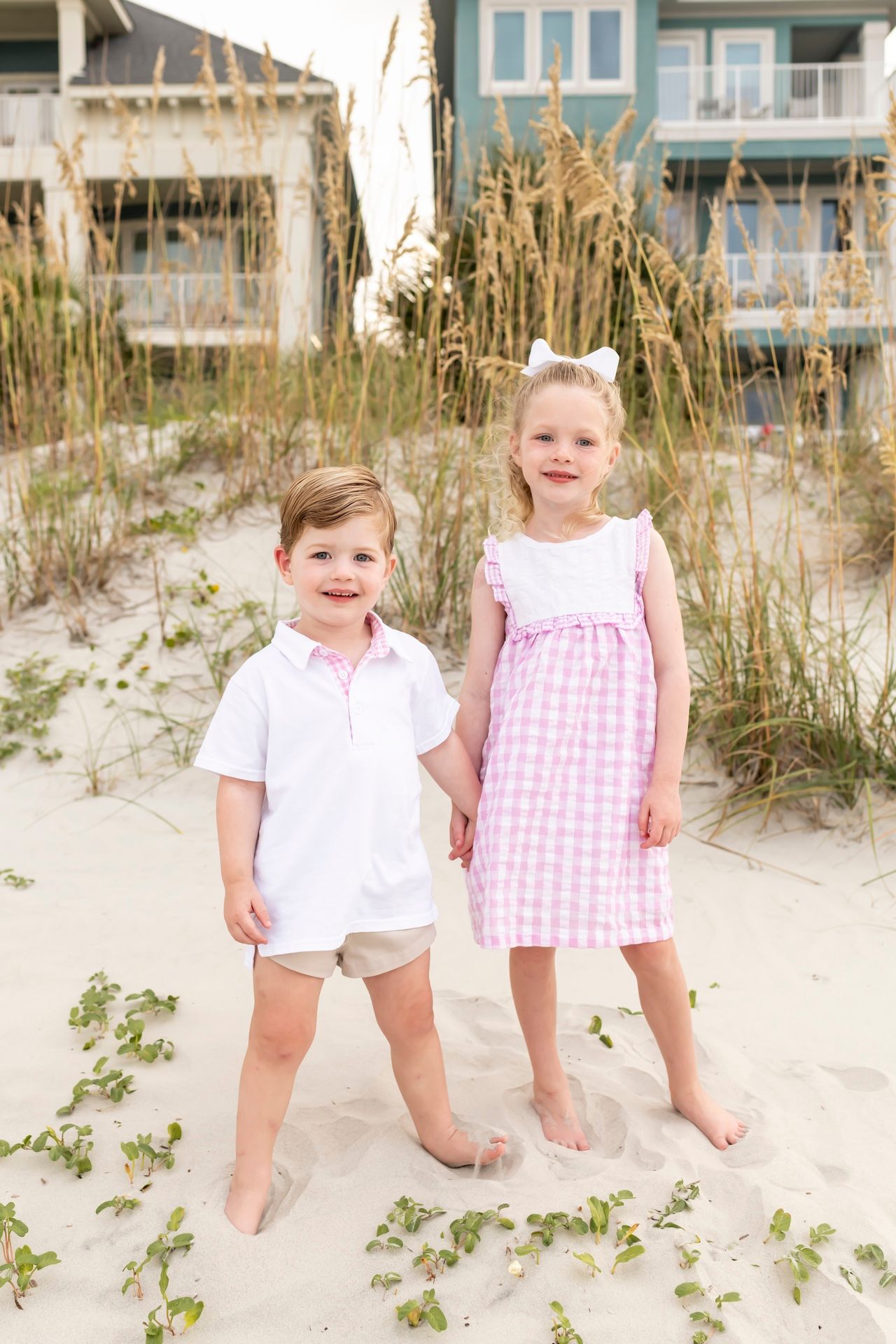 Young siblings wearing white and pink gingham clothing pose together on a sandy beach with beach houses behind them.