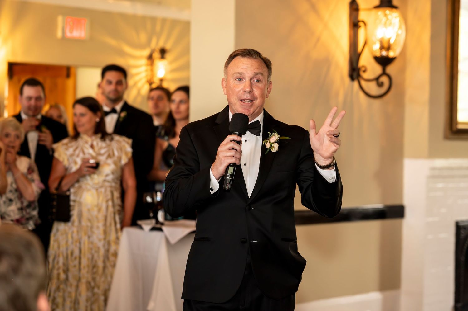 A man in a black tuxedo gives a speech at an elegant event while guests listen in the background.