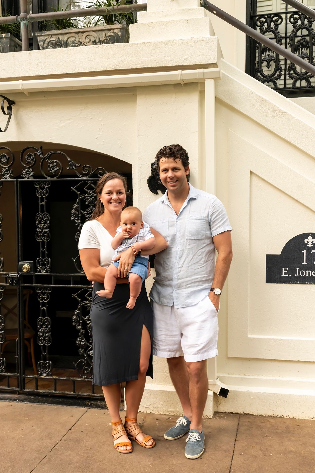 A family stands together outside a cream-colored townhouse with ornate black iron railings and architectural details.