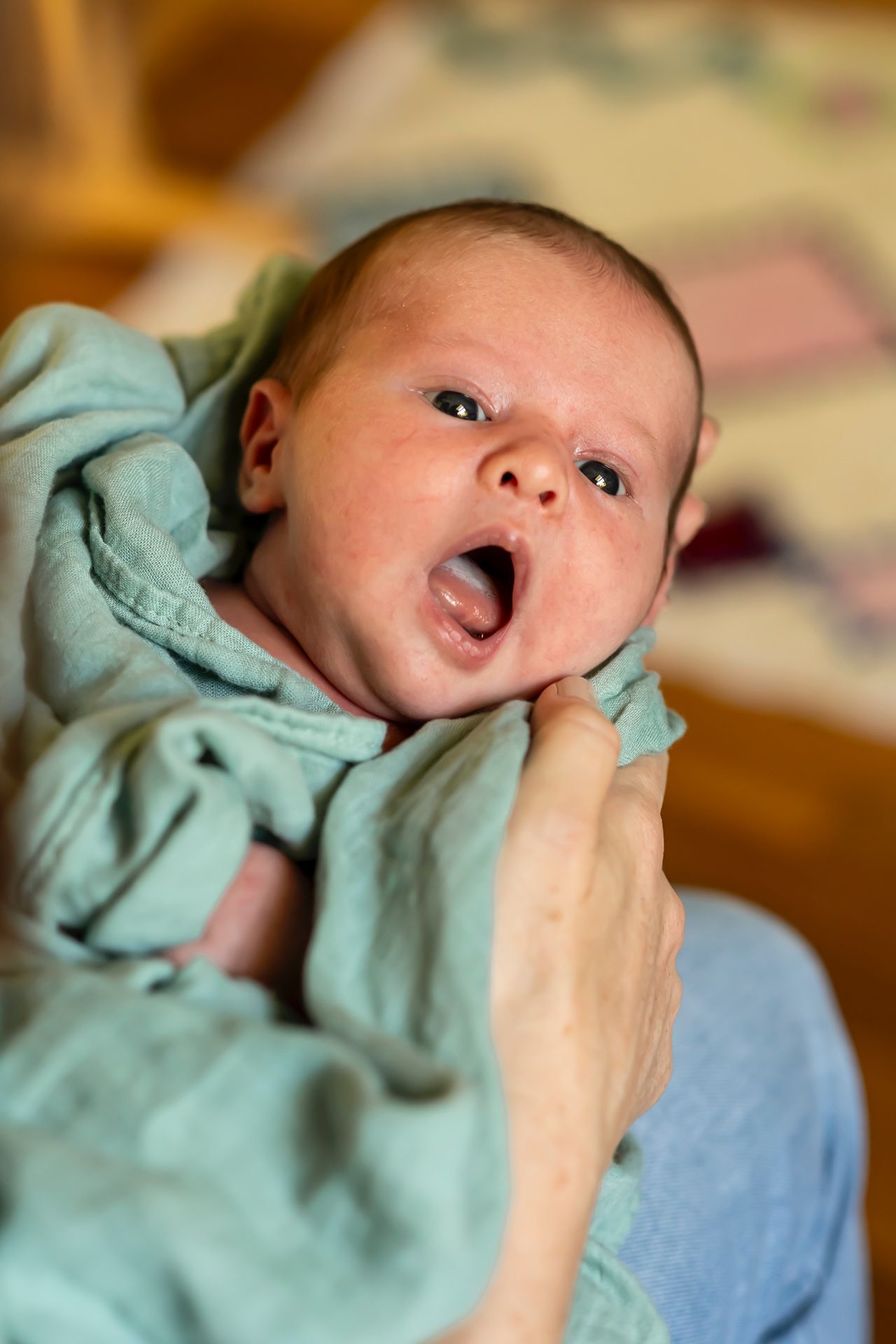 A baby in a mint green outfit yawns while being held, photographed in warm lighting from above.
