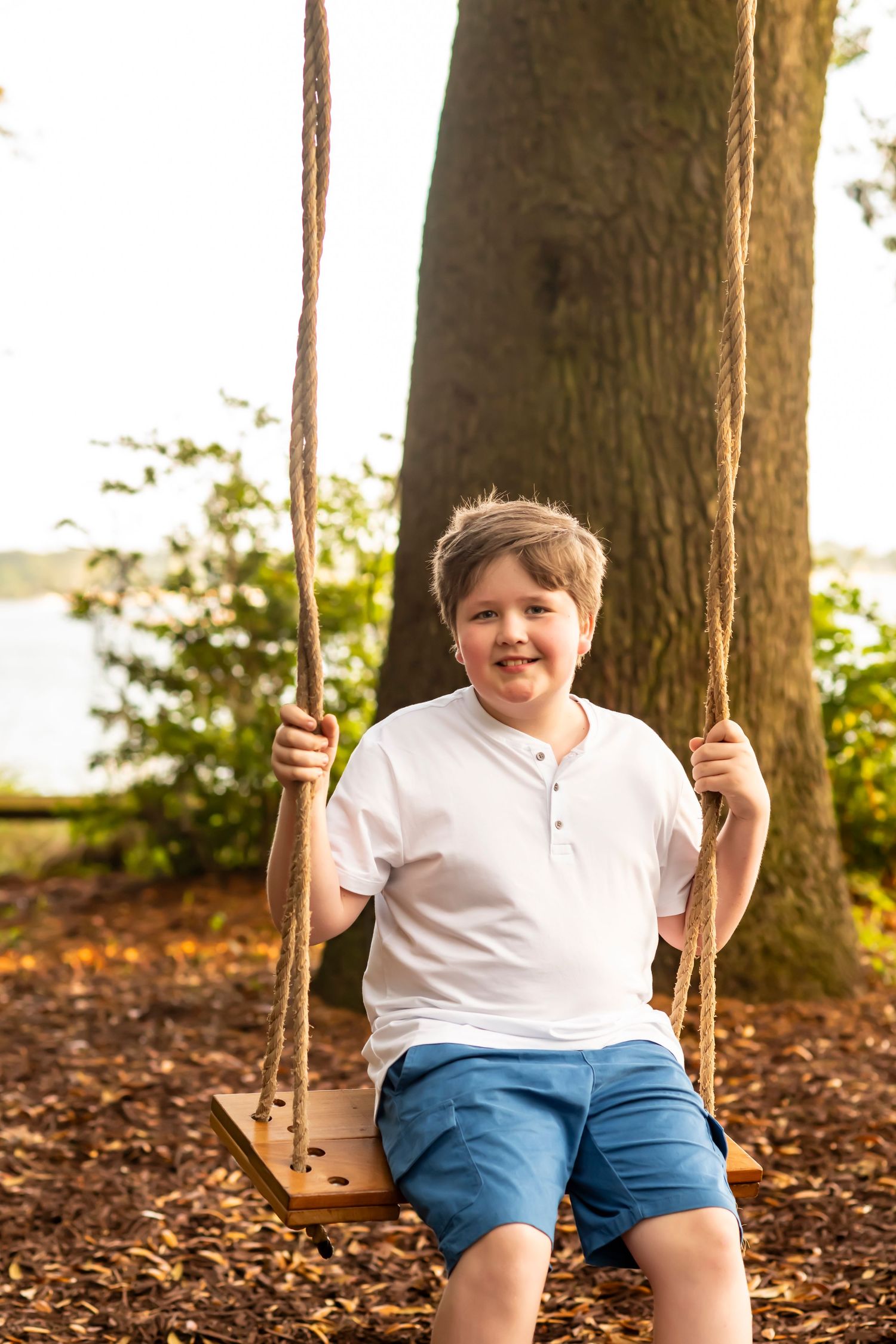 Young child smiling while sitting on a wooden rope swing under tall trees at sunset by a lake.
