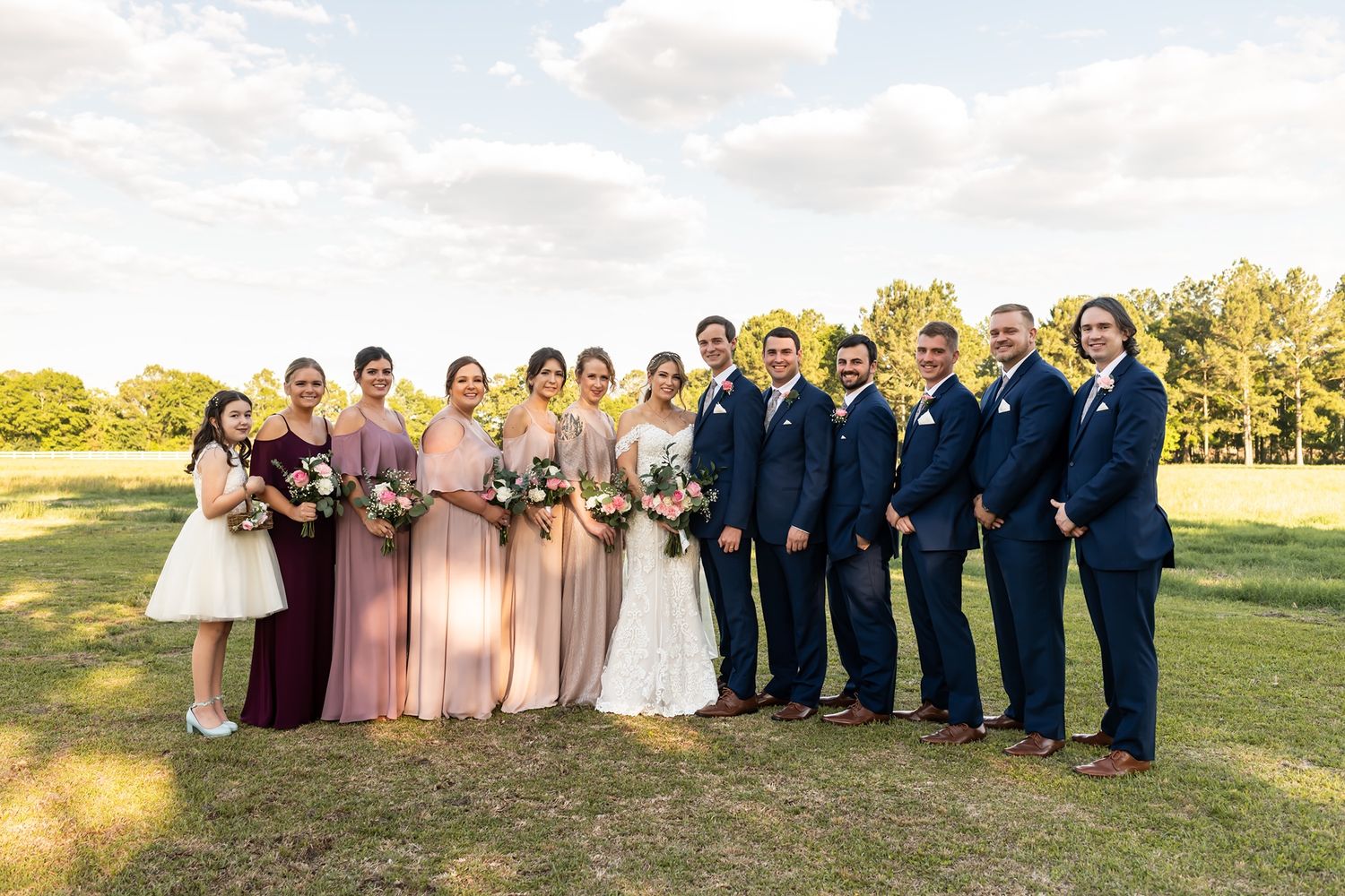 A wedding party in formal attire poses outdoors on a sunny day with bridesmaids in pink and groomsmen in navy suits.